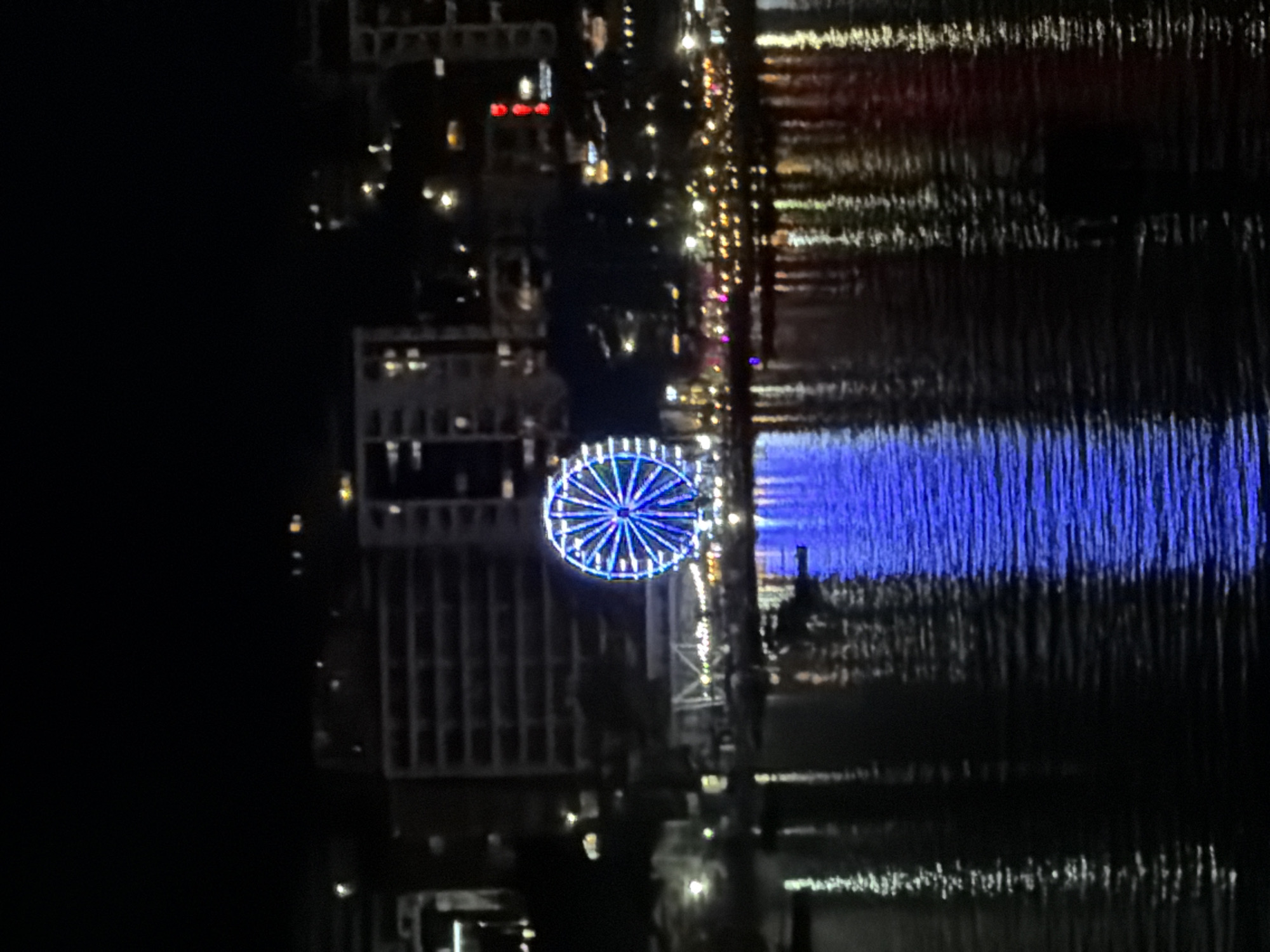 The image captures a nighttime view of a Ferris wheel illuminated with blue lights, prominently situated in the city of Boreti, Montenegro. The Ferris wheel is reflected in the dark, rippling water of the bay in front of it. In the background, several multi-story buildings are visible, some with illuminated windows, suggesting an urban environment. The overall scene is dark, with the bright lights of the Ferris wheel and buildings creating points of illumination against the night sky and water. There are no visible people or any specific actions taking place, beyond the static presence of the Ferris wheel and surrounding structures. The water surface shows distinct reflections of the lights from the Ferris wheel and the city.