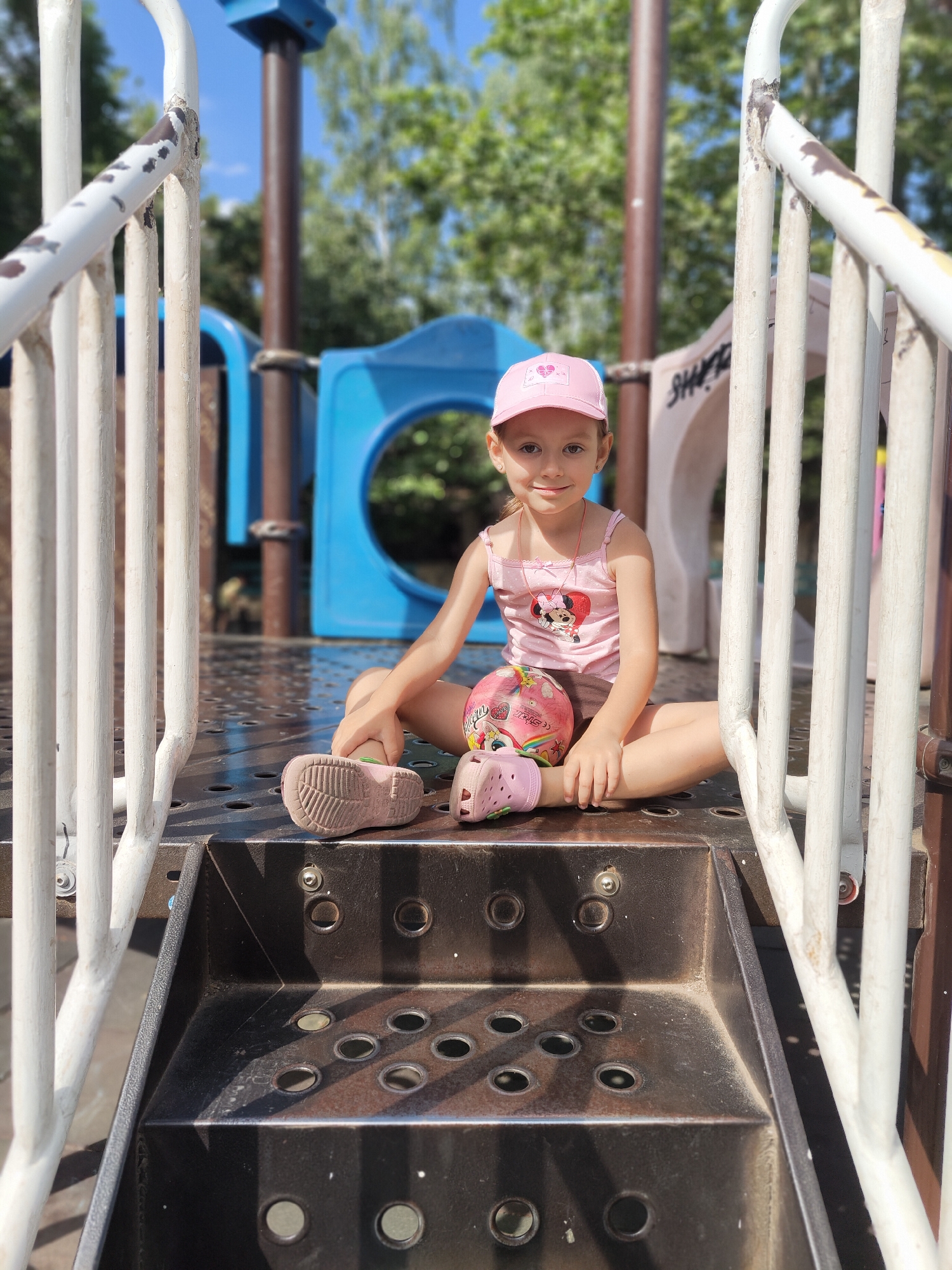 A young girl with a pink baseball cap and a pink tank top with Minnie Mouse on it sits on the steps of a playground in Chornomorsk, Ukraine. She is holding a pink ball with cartoon characters on it. The playground equipment is made of metal and plastic. Trees and a blue sky are visible in the background. The girl is smiling at the camera. She is wearing pink Crocs. The photo was taken in daylight.