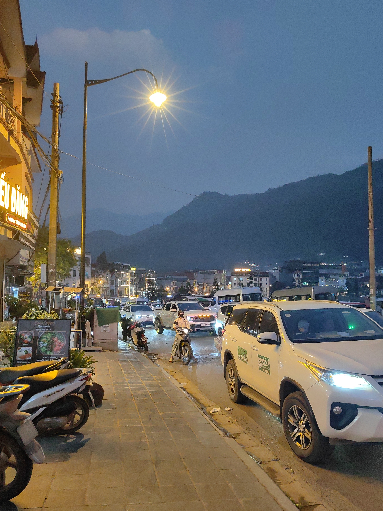 The image presents a street scene in Sa Pa, Vietnam, captured at dusk or early night. The sky is a deep blue-gray with subtle cloud formations, and various artificial light sources illuminate the environment.
In the foreground on the left, a paved sidewalk features three parked motorbikes; a white scooter is most prominent, followed by two darker models. Immediately adjacent, a black, vertically oriented signboard advertises a "Restaurant" with food graphics and contact information in Vietnamese. Behind this, green vegetation is visible. A multi-story building with a yellow exterior occupies the left side of the frame, displaying a large, brightly illuminated sign with Vietnamese text "ĐIỆU BẰNG" and a phone number (0983 998 298). A utility pole densely packed with wires stands in front of this building.
The asphalt road dominates the mid-ground, bustling with vehicular traffic. A street light positioned centrally over the road emits a strong, starburst-effect glow. A person on a white motorbike, dressed in a light-colored shirt, is driving towards the left side of the frame. Behind this motorbike, a white pickup truck, likely a Ford Ranger, is visible with its headlights activated, positioned within a line of slow-moving or stopped vehicles. Further down the street, additional cars and a white passenger van or bus are discernable amidst the illuminated facades of urban buildings.
On the right side of the road, a white Toyota Fortuner SUV is positioned, displaying "TAXI XANH" decals on its side, indicating its function as a taxi. Its headlights are on, and a passenger, possibly a child, is visible in the front seat.
The background reveals a dense cluster of buildings with numerous illuminated windows and signs, including one explicitly marked "REY HOTEL." A dark, formidable mountain range rises prominently in the distant background, its contours starkly silhouetted against the fading light of the sky and the ascending urban glow of Sa Pa.