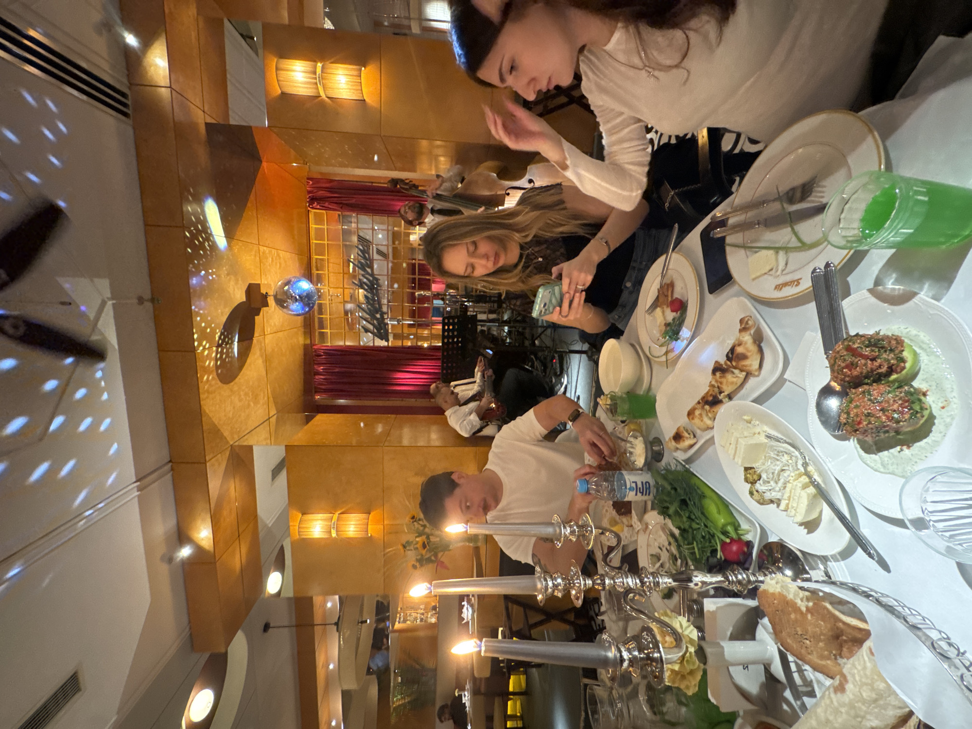 The image captures a dining scene in what appears to be a restaurant in Yerevan, Armenia. Three individuals are seated around a table laden with food. Two women are visible on one side of the table; one is looking at her phone, while the other seems to be adjusting her hair. A man is seated on the other side of the table, reaching for something amongst the food. The table is covered with various dishes, including bread, salads, and pastries. There are also several bottles and glasses on the table. In the background, a live band consisting of a guitarist and a cellist is performing behind a curtain. The interior decor of the restaurant includes warm lighting, wooden panels, and a disco ball hanging from the ceiling.