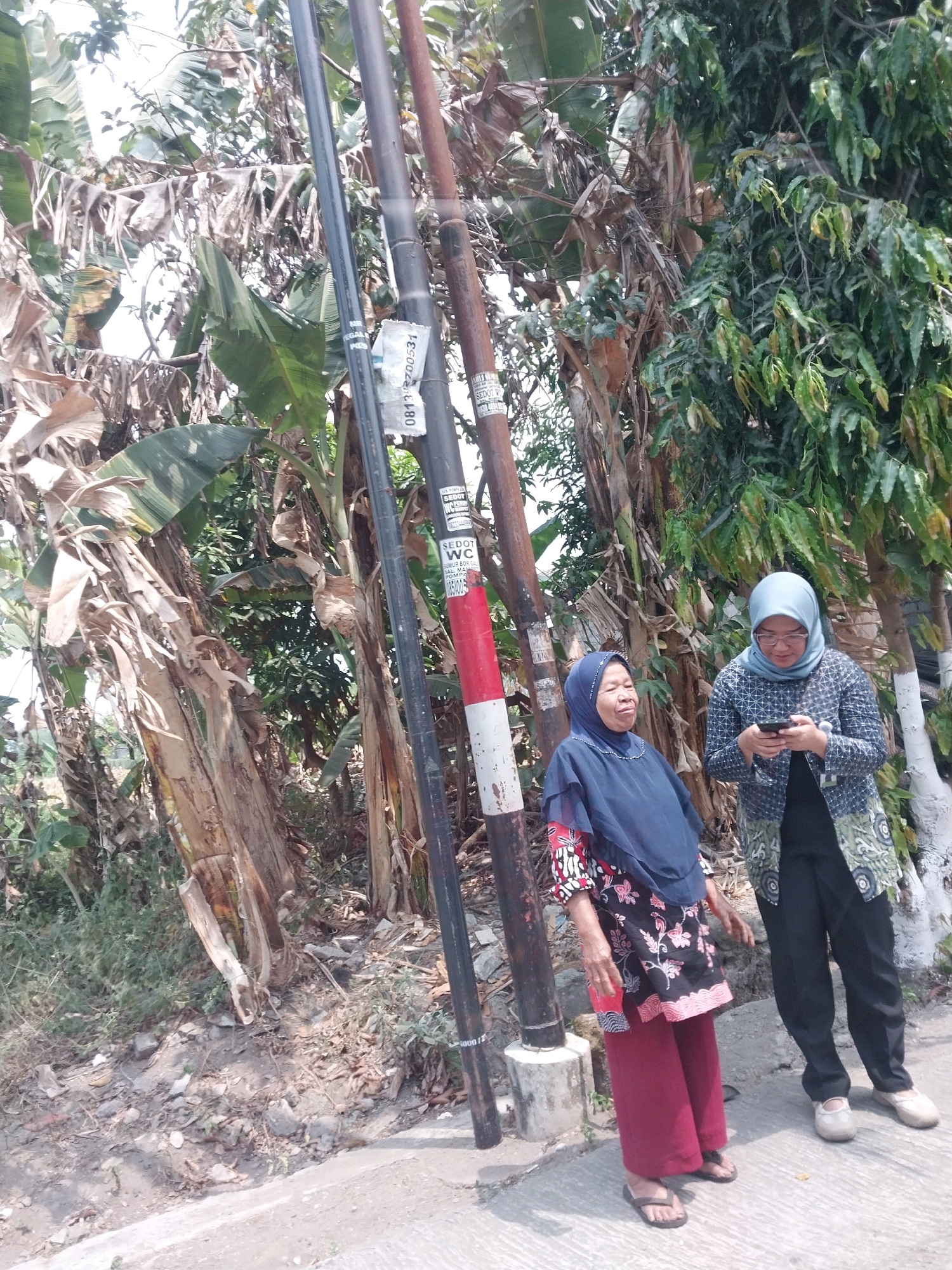 In the city of Tirtomartani, Indonesia, two women stand on a paved surface next to a pole with red and white markings and another utility pole. The woman on the left, wearing a dark blue hijab and a patterned top with red sleeves, looks to her left. The woman on the right, wearing a light blue hijab and a patterned jacket over dark pants, is looking down at her smartphone. The background consists of lush vegetation, including banana trees and other plants, with dry leaves scattered on the ground. The lighting suggests it is daytime, with shadows indicating sunlight.