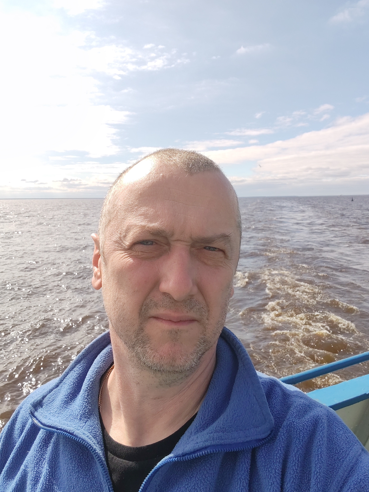 A middle-aged, Caucasian male with short, graying hair is taking a selfie. He is wearing a blue fleece jacket and a black shirt underneath. A thin gold chain is visible around his neck. He has light blue eyes and a stubbled beard. The background shows a body of water, possibly a river or a lake, with choppy waves. The sky is partially cloudy. Based on the clues, the location could be related to the city of Mologa, Russia, before it was flooded and submerged, or a present-day location that resembles the submerged region. However, there is no concrete information to confirm the exact location in the image. The scene suggests a casual moment captured on a boat or near a body of water.