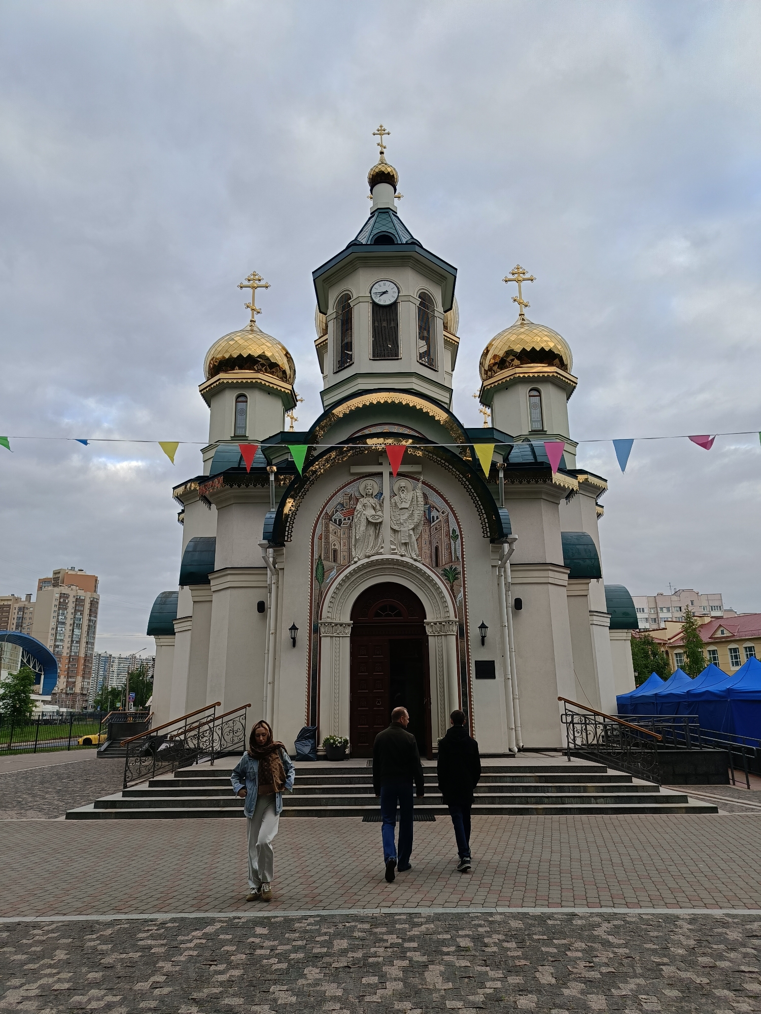The image shows a church, likely located in Saint Petersburg, Russia. The church is a white building with three golden domes topped with Orthodox crosses. The central dome is taller and includes a clock. The entrance to the church is adorned with religious figures in mosaic or relief. Strings of colorful triangular flags are strung across the front of the church. Three people are walking toward the church: a woman wearing a denim jacket and light-colored pants is in the foreground, while two men are slightly behind her. The ground is paved with patterned brick. In the background, there are apartment buildings and a blue tent. The sky is overcast.