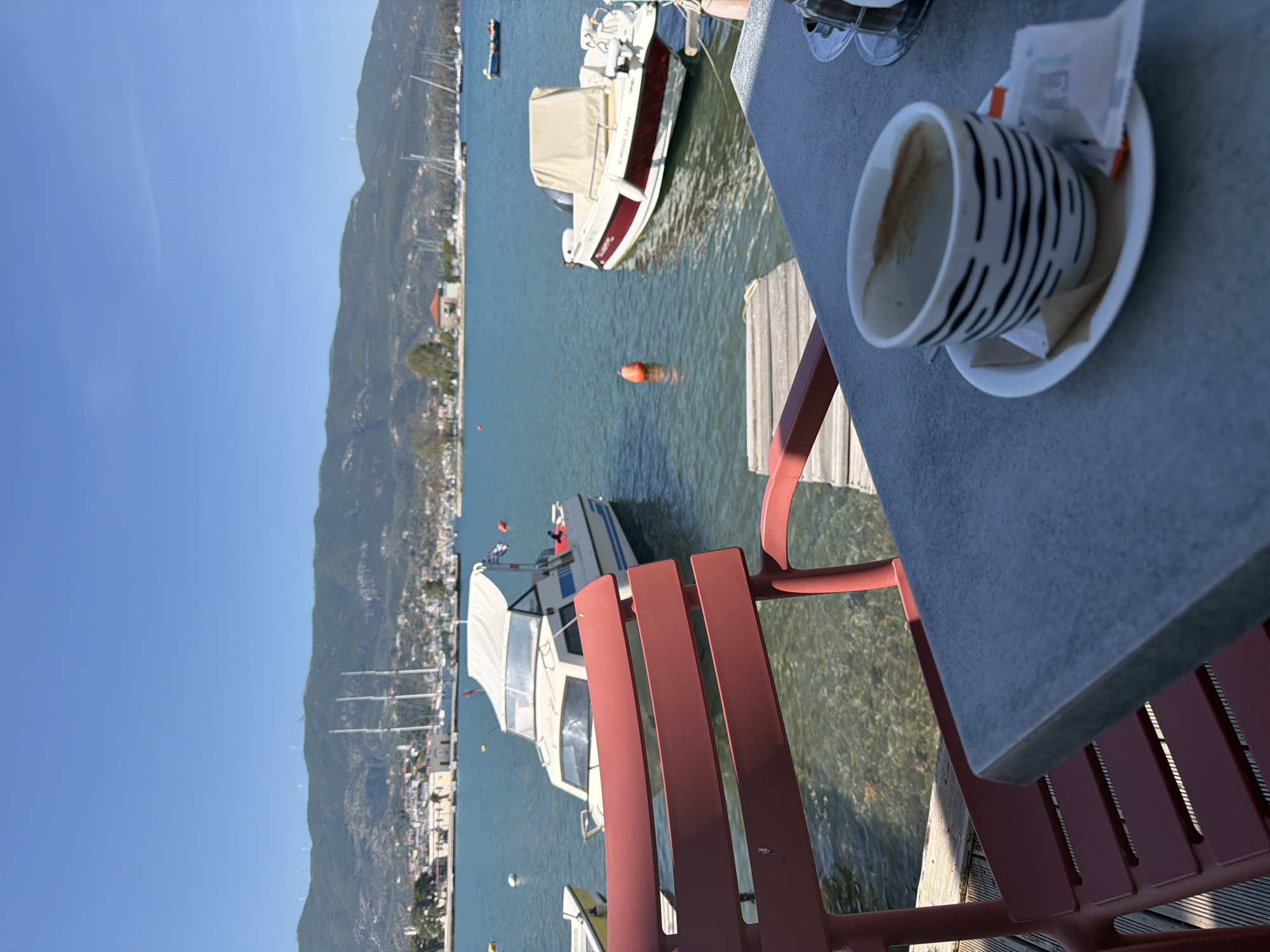 The image captures a serene waterfront scene, seemingly from a cafe or restaurant terrace, rotated 90 degrees clockwise. In the immediate foreground, a textured gray tabletop is visible, holding a white and black striped coffee mug, partially empty, on a white saucer. Next to the mug are small paper packets, one of which appears to be sugar, with faint text including "Love Coffee" visible. A section of a red outdoor chair, made of slats, is prominent in the lower right, positioned next to the table.
Beyond the terrace, clear, calm blue-green water extends, revealing the seabed in shallower areas. Several boats are present on the water. The closest is a white motorboat with a red stripe along its hull and a beige canvas cover, seen from its stern. An orange buoy floats in the water near this boat. Further in the distance, other boats, including several with prominent masts suggesting sailboats, are moored along the coastline.
The background features a long, verdant mountain range running along the horizon, dotted with buildings and what appears to be a small coastal town or village. The sky above is a bright, clear blue, indicating sunny weather. The overall atmosphere suggests a leisurely moment on a bright day by the sea, likely in a Mediterranean setting such as Poros, Greece.