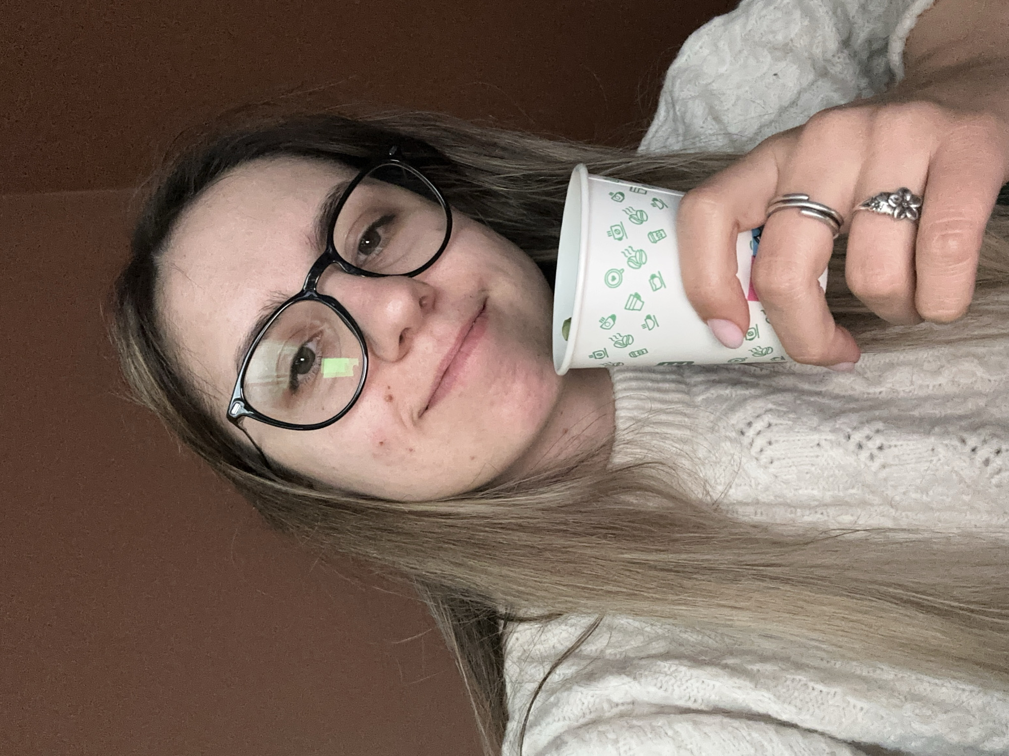A young woman with brown hair and glasses poses for a selfie. She's holding a white disposable cup with green food-related icons, adorned with two silver rings on her fingers. She is wearing a white sweater. The background is a plain brown wall. The image does not provide enough information to determine a location in Sofia, Bulgaria.