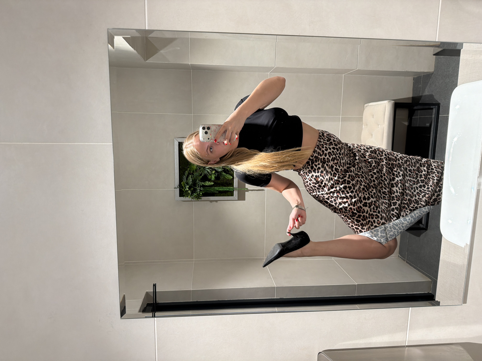 A woman is taking a mirror selfie in a bathroom. She is wearing a black crop top and a leopard-print skirt with black high heels. The bathroom has light gray tiled walls and a mirror with a thin black frame. A small framed plant decoration is visible on the wall behind her. A padded stool and a dark cabinet are also visible in the background. The location in the city of Izhevsk, Russia cannot be determined.
