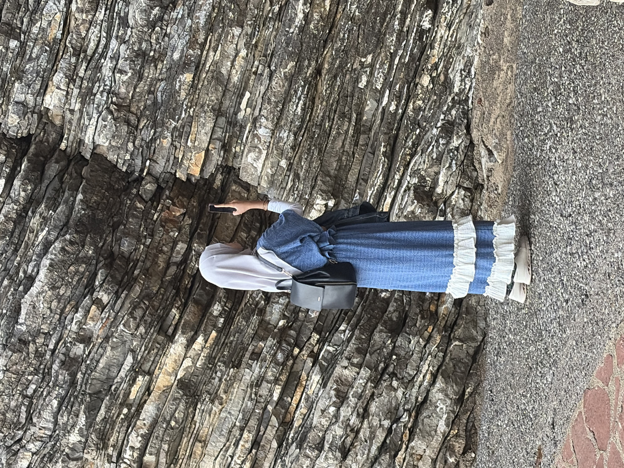 A woman in a long, blue dress with white ruffles at the hem and a gray headscarf is standing next to a large rock formation. She is holding a smartphone in her hand, possibly taking a photograph. The woman has a black purse slung over her shoulder. The ground to the right of the rock formation appears to be a paved area with small gravel. The location is likely in or near Budva, Montenegro, given the rocky coastline and urban environment. The woman seems to be interacting with her environment by capturing a picture of the rock formation.