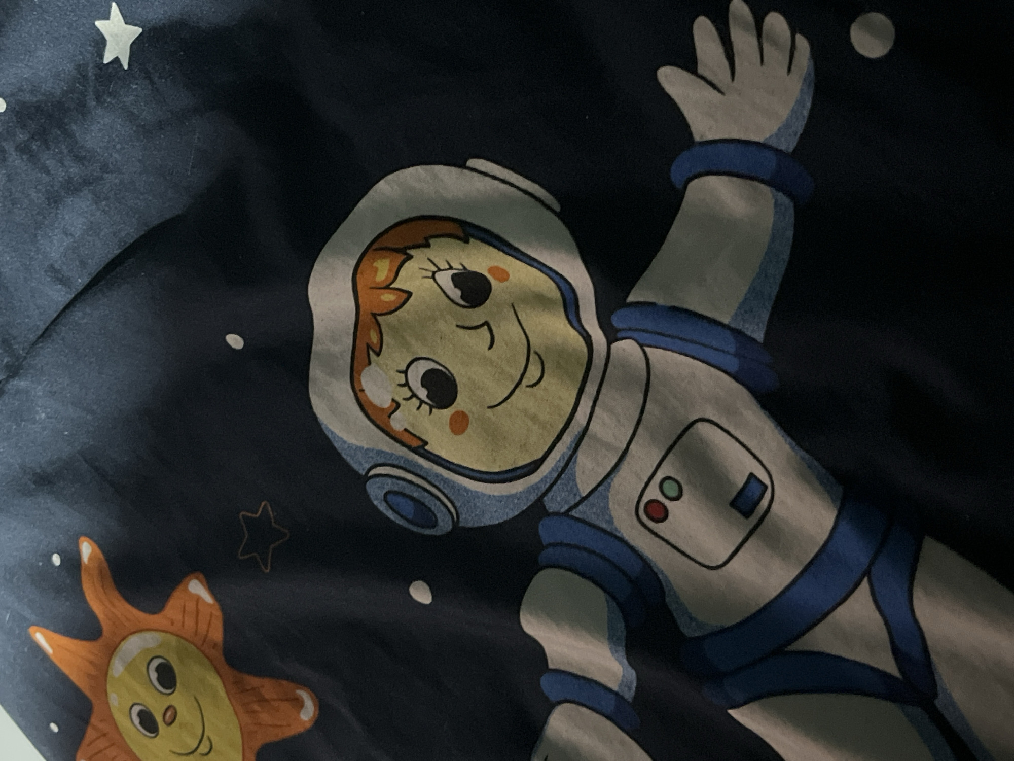 This is a close-up, overhead shot of a dark blue fabric with a children's space-themed print. The print features a cartoon astronaut in a white and blue suit, with a light yellow face and orange hair. The astronaut is depicted with a smiling expression, open eyes, and eyelashes. The suit has a helmet with a blue visor, blue accents on the sleeves and legs, and a control panel on the chest with red, green, and blue buttons. To the upper left of the astronaut is a cartoon sun with a smiling face, orange rays, and lighter yellow highlights. The background of the fabric is dark blue and scattered with white dots and a faint outline of a star. The fabric appears to be a sheet or pillowcase, and it is folded or bunched in places, creating shadows and highlights that obscure some details. The image was taken in Denasri Kulon, Indonesia.