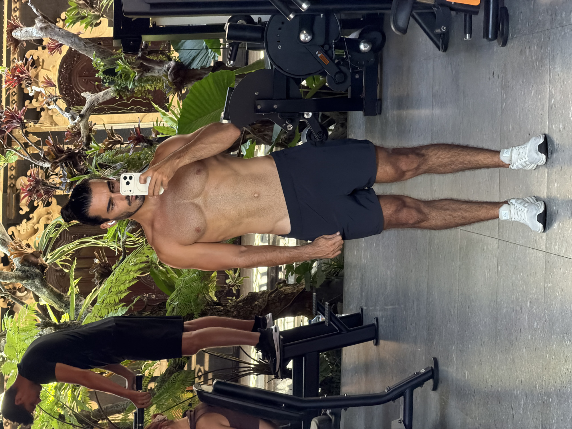 The image shows a shirtless man taking a selfie in a gym in North Kuta, Indonesia. He has dark hair pulled back in a bun and is wearing dark shorts and white athletic shoes. He stands next to an exercise machine. The gym is open to the outdoors and features a tropical garden with ferns and various plants. Another person is visible in the bottom left corner, also in gym attire, operating an exercise machine. The floor is a light grey, speckled tile.