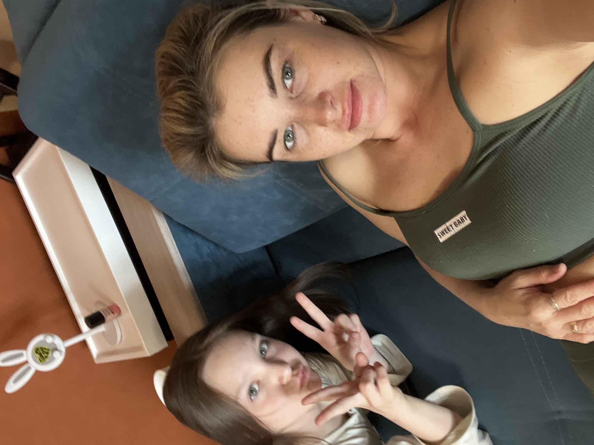 Two people are lying on a blue sofa. The person on top is an adult female with fair skin, blonde hair pulled back in a bun, freckles, and wearing a dark green tank top with a small white label that reads "SWEET BABY". She is looking at the camera.
The person below is a child with fair skin and long brown hair, making a peace sign with both hands. They are wearing a light-colored top.
Next to the sofa is a white and black rectangular object with a small item resembling a bunny attached to its side. The floor is brown.
It is not possible to confirm that the location is Chaikovsky, Russia based on the image.