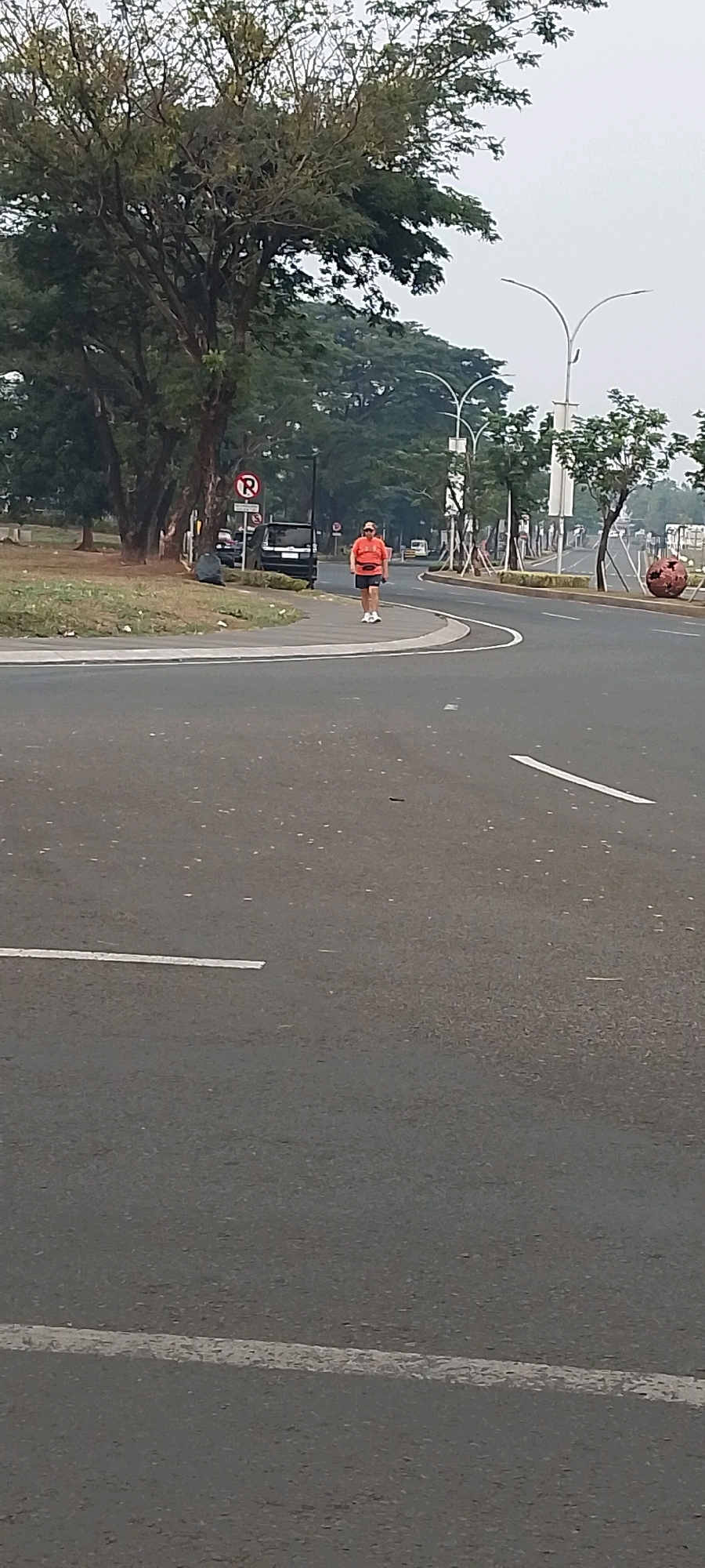A person is jogging on the sidewalk adjacent to a road in Gunung Putri, Indonesia. The jogger is wearing an orange t-shirt and dark shorts, and appears to be carrying a small orange pouch around their waist. A dark-colored SUV is parked on the sidewalk to the left of the jogger, next to a circular sign with a red "P" crossed out. The road has a white line marking its lanes, and several streetlights are visible in the background. Trees with green foliage line the side of the road, and the sky is overcast. The overall scene depicts an outdoor recreational activity in a suburban or park-like setting within the city.