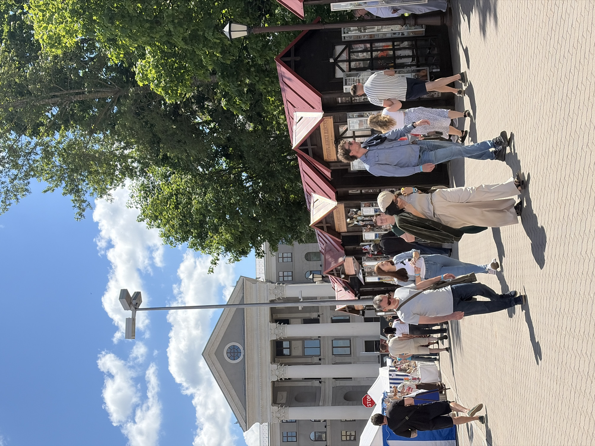 The image shows an outdoor market scene in Minsk, Belarus, set against the backdrop of a classical building with prominent columns. The sky is blue with scattered white clouds. A large tree with lush green foliage is visible on the left side of the image.
A row of market stalls with red roofs lines the walkway, with people browsing and interacting with the vendors. Individuals of various ages and appearances are present, some walking, others standing at the stalls. Clothing styles vary, indicating a casual atmosphere.
A light post is located near the building. The ground is paved with light-colored tiles. A "STOP" sign is visible.