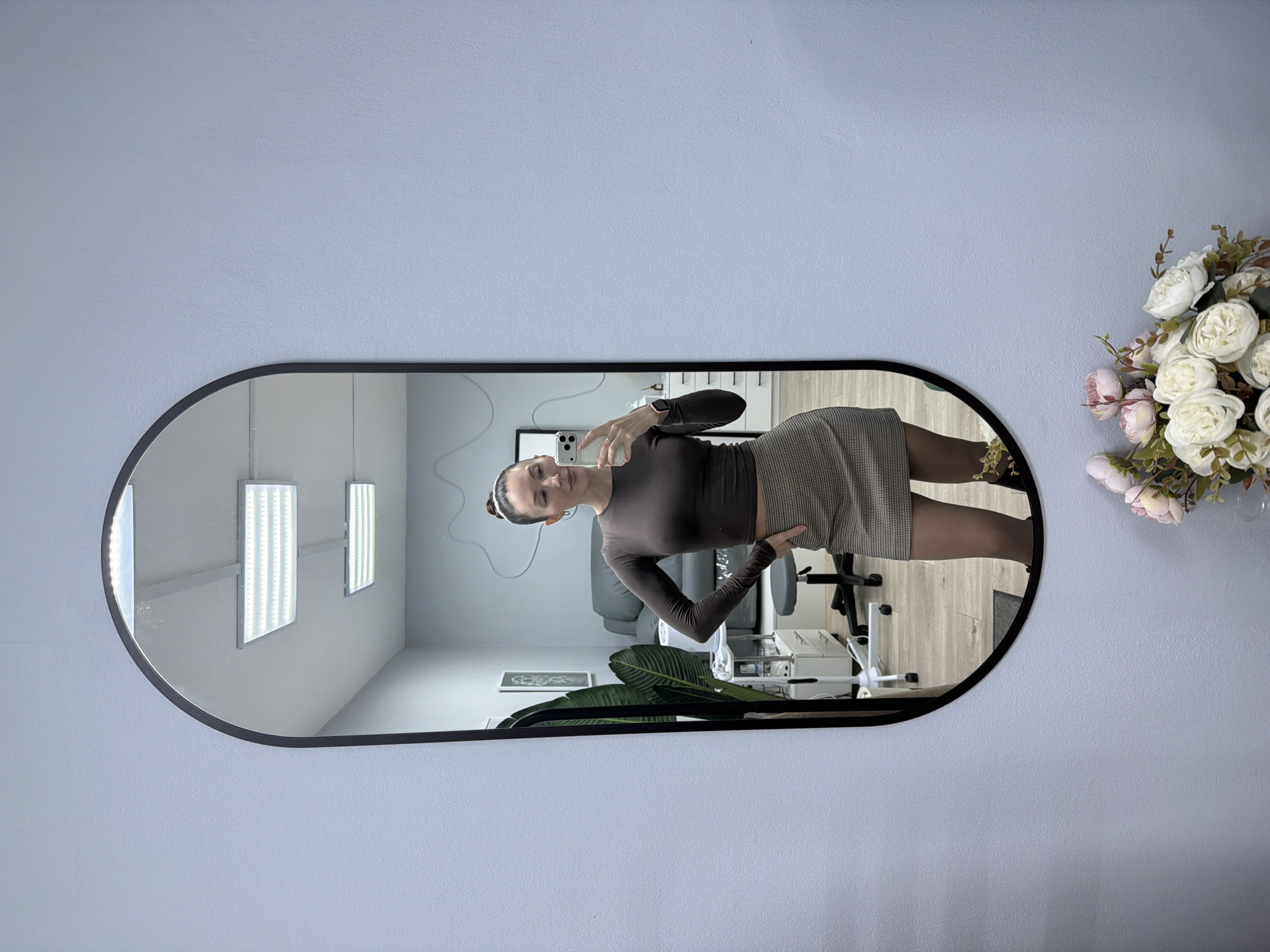 A young woman is taking a mirror selfie. She is indoors in a room in Perm, Russia, that appears to be a salon or spa. She is wearing a long-sleeved brown shirt, a tan skirt, and dark stockings. In the background is a salon chair, a table with equipment, and decorative plants. The wall is light blue, and a bouquet of white and pink flowers is in the upper right corner of the frame. The mirror has a rounded rectangular shape with a black frame.