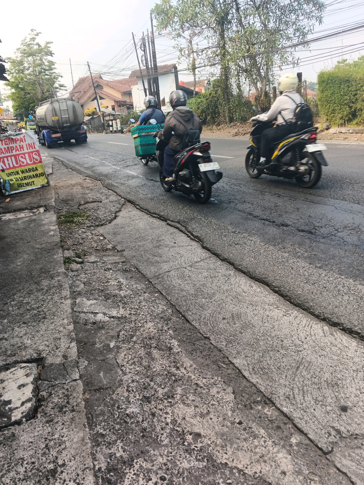 The image captures a street scene in Ungaran, Indonesia, likely on a cloudy day. In the foreground, a cracked and uneven concrete sidewalk runs along the left side of the frame, with some sparse vegetation growing through the cracks. The road surface is asphalt and appears wet, with visible cracks and patches.
Several vehicles are present on the road. On the left, a large silver tanker truck is moving away from the viewer. To its right, two motorcycles are traveling in the same direction. The first motorcycle has a basket attached to its rear, and the rider is wearing a blue shirt and a helmet. The second motorcycle is positioned slightly behind and to the right of the first, with a rider wearing a white shirt and a backpack, also with a helmet. Another motorcycle, black with a white license plate, is partially visible in the lower left foreground.
The background features buildings with terracotta roofs and trees lining the street, with power lines overhead. A red and yellow sign is visible on the left side of the road, with text in Indonesian that appears to be a notice or announcement. The overall impression is of a busy but somewhat weathered urban environment.