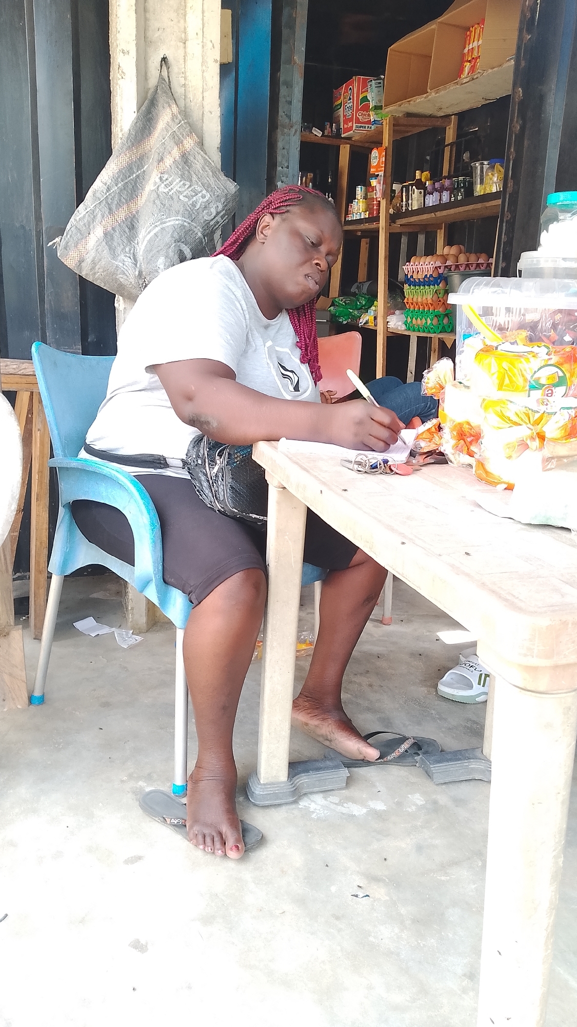 A woman sits at a table outdoors, writing on a piece of paper. She has red braids and is wearing a gray t-shirt and black shorts. A black fanny pack is slung across her waist. The table has items on it, including a container with snacks. She is sitting on a blue plastic chair, and her bare feet rest on top of her sandals and a small piece of gray plastic. The floor is concrete. In the background, there's a shelf stocked with food items, and a cloth sack is hanging from a blue and black metal frame.