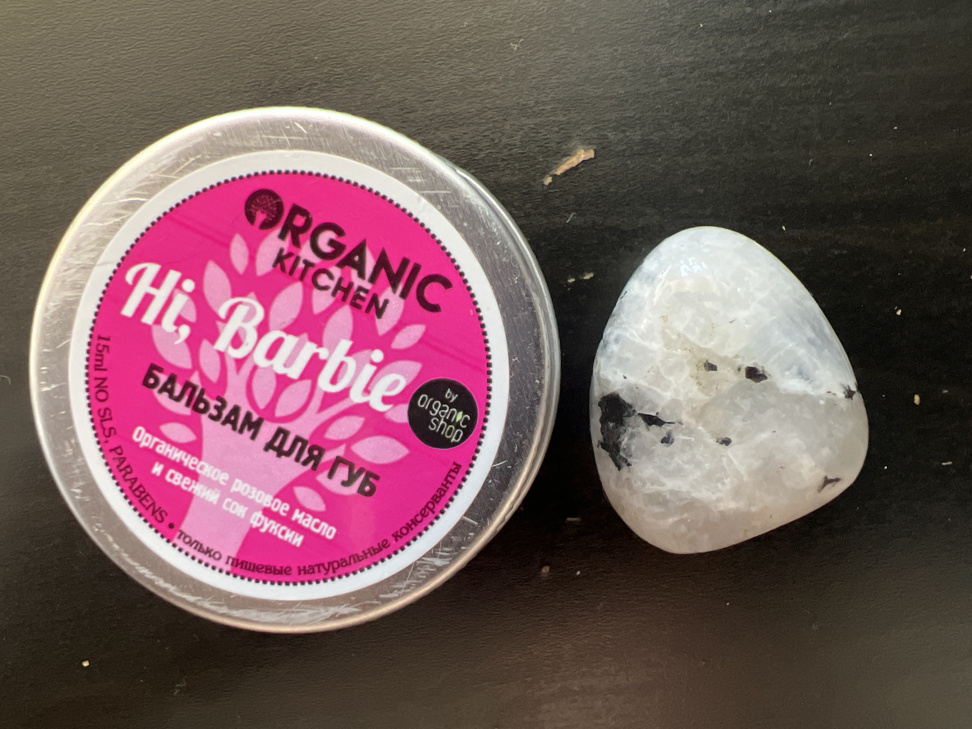 A close-up, top-down shot captures a pink and silver tin of "Hi, Barbie" organic lip balm resting on a dark, textured surface. The tin has circular branding with floral patterns and Russian text. Below the tin, a smooth, white, and gray stone with black inclusions lies on the surface. The scene appears to be indoors, as indicated by the lighting and indoor textures. No specific city or geographical markers are visible in the image.