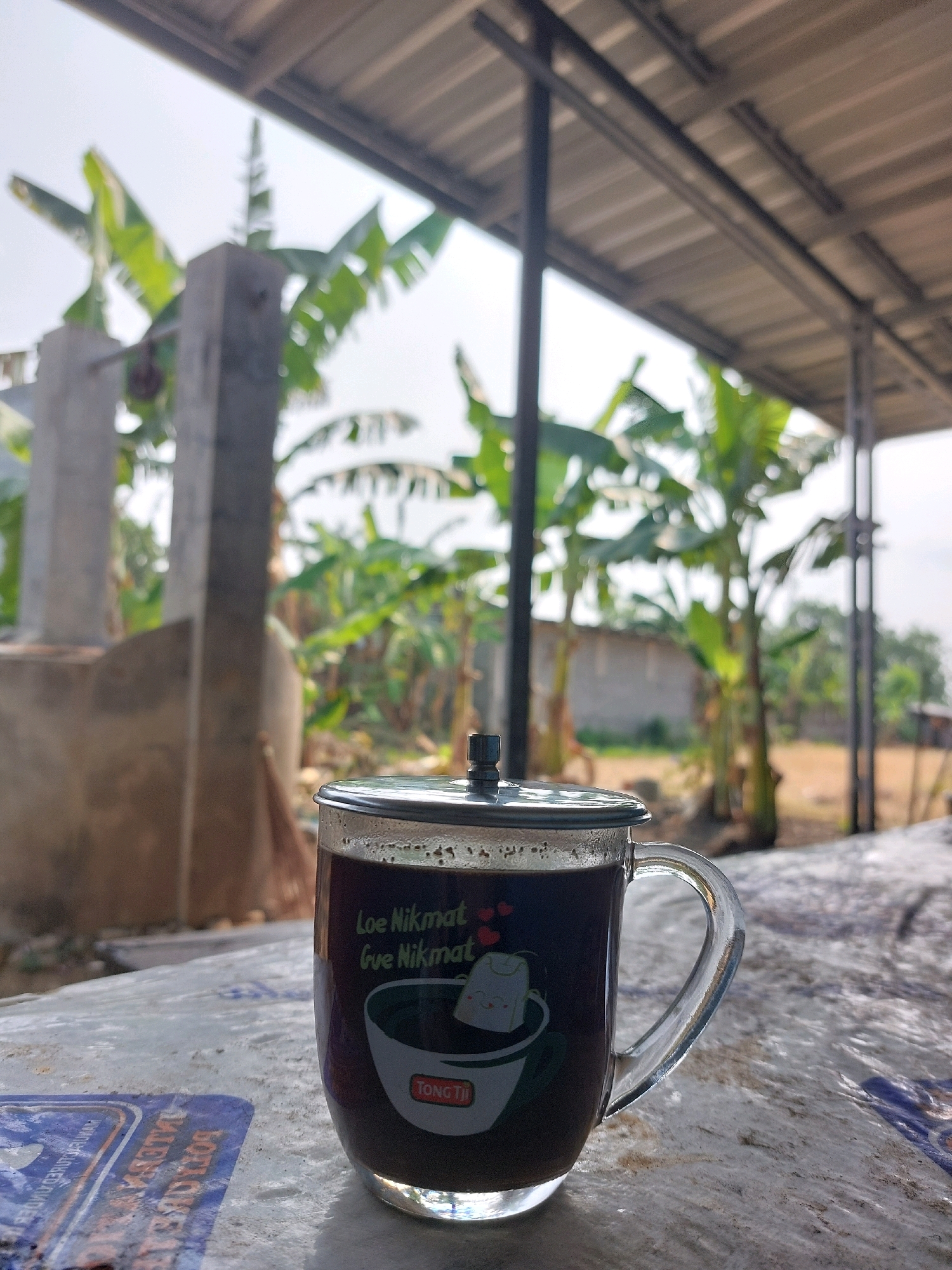 A close-up, low-angle shot captures a glass mug filled with dark liquid, likely coffee, resting on a textured surface. The mug features a lid and a handle, with a design on its side that appears to depict a teabag in a cup, accompanied by text that reads "Loe Nikmat Gue Nikmat". The background is blurred, showing lush green banana trees and a rustic structure, suggesting an outdoor setting. The overall ambiance is casual and relaxed, possibly in a rural or semi-rural environment, such as a backyard or a simple eatery in Wonokarto, Indonesia.