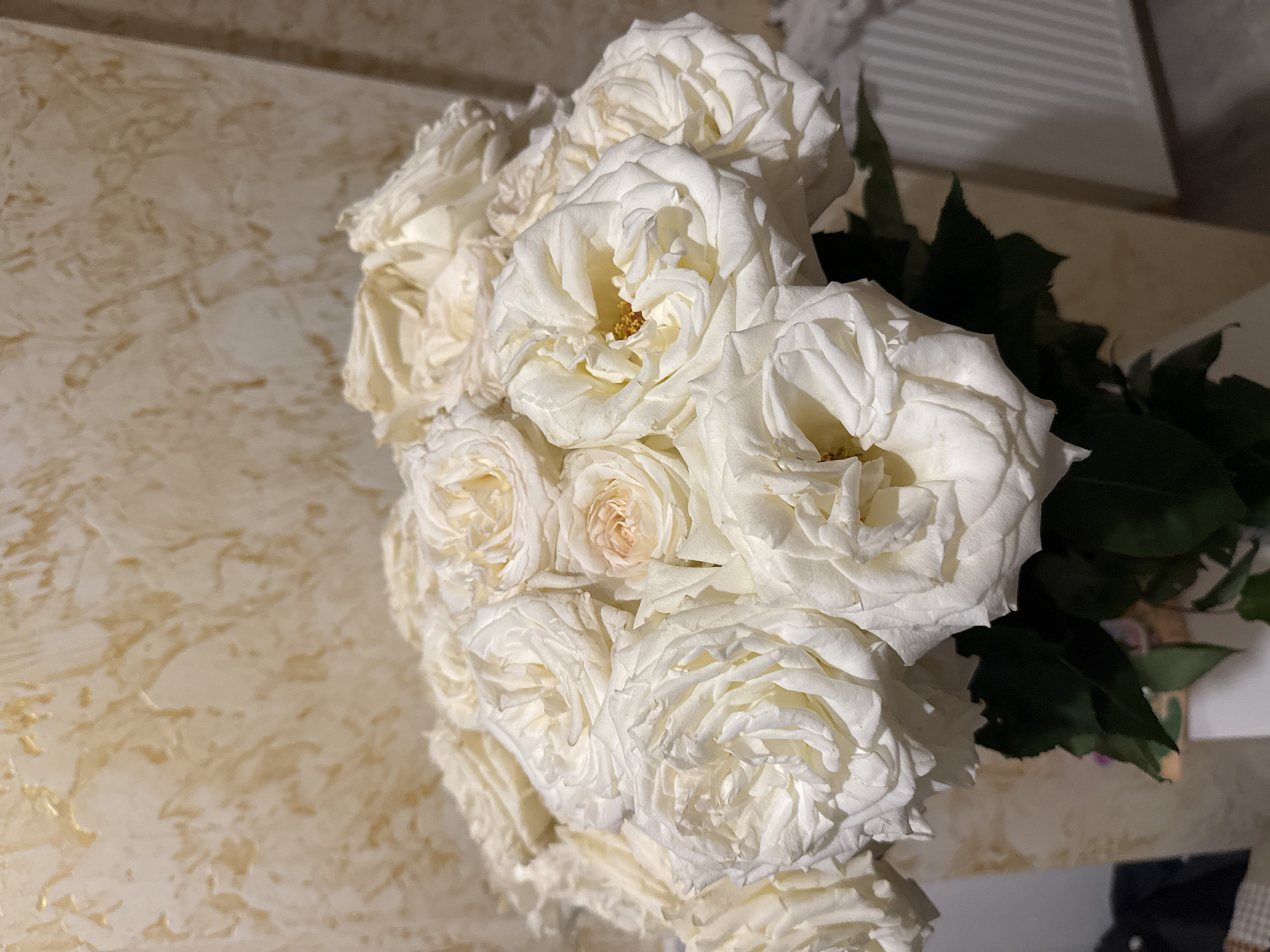 A cluster of white roses with yellow and peach centers is positioned on a surface marked with a swirling pattern. Green leaves emerge from behind the roses. The scene is illuminated by an unseen light source.