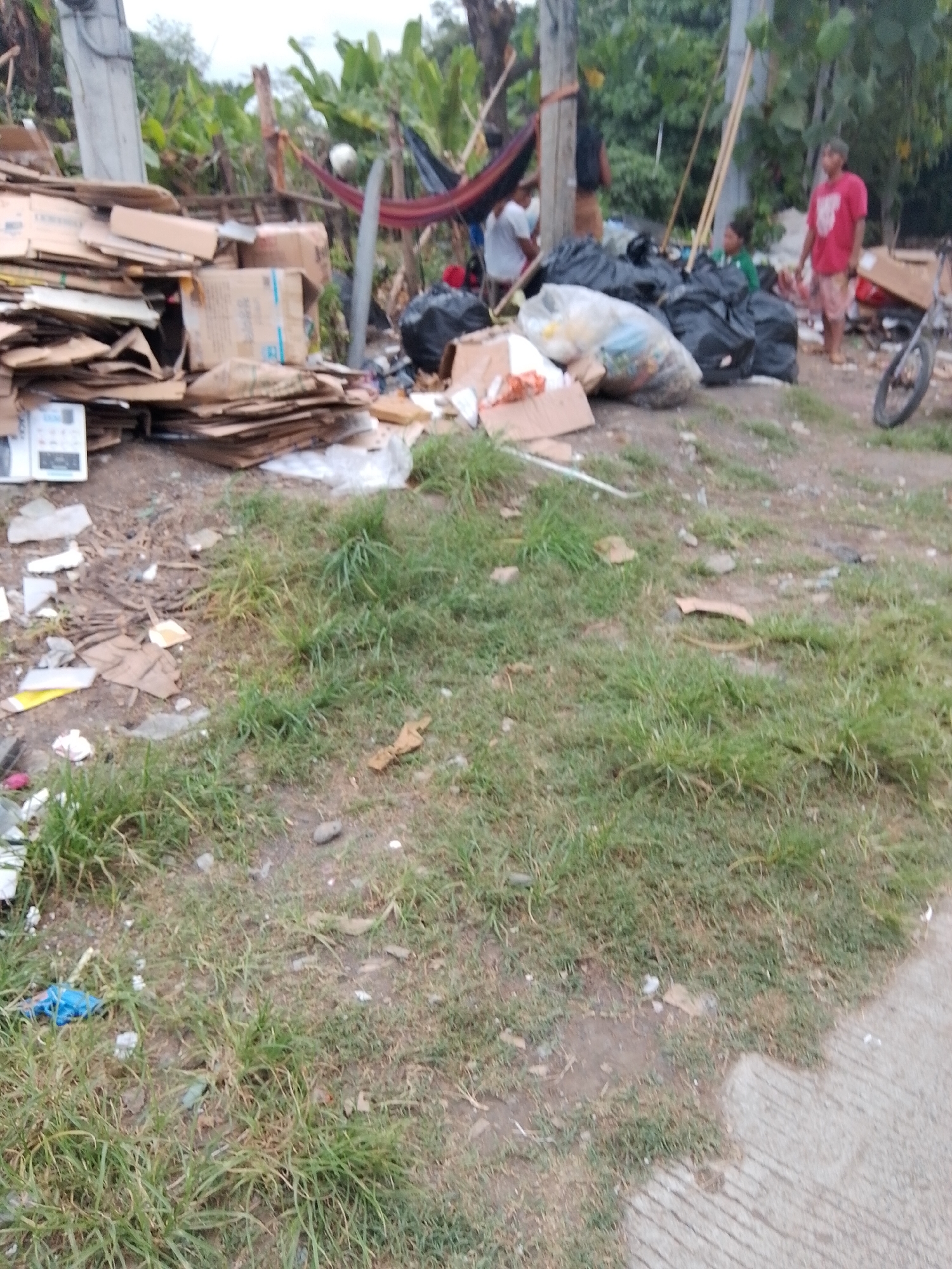 The image depicts an outdoor scene in Davao City, Philippines, characterized by a significant amount of debris and discarded materials. In the background, several individuals are visible near a hammock strung between two poles, suggesting a makeshift living or resting area. One person is wearing a red t-shirt and light-colored shorts. A pile of large black trash bags dominates the middle ground, alongside a clear plastic bag filled with various items, possibly recyclable materials. To the left, a large stack of flattened cardboard boxes and other packaging materials is piled up. Various pieces of trash, including paper, plastic, and debris, are scattered across the ground, which is covered with patchy grass and dirt. A section of a concrete path is visible in the lower right corner. The overall impression is one of disarray and waste accumulation, potentially in an informal settlement or an area designated for waste disposal.