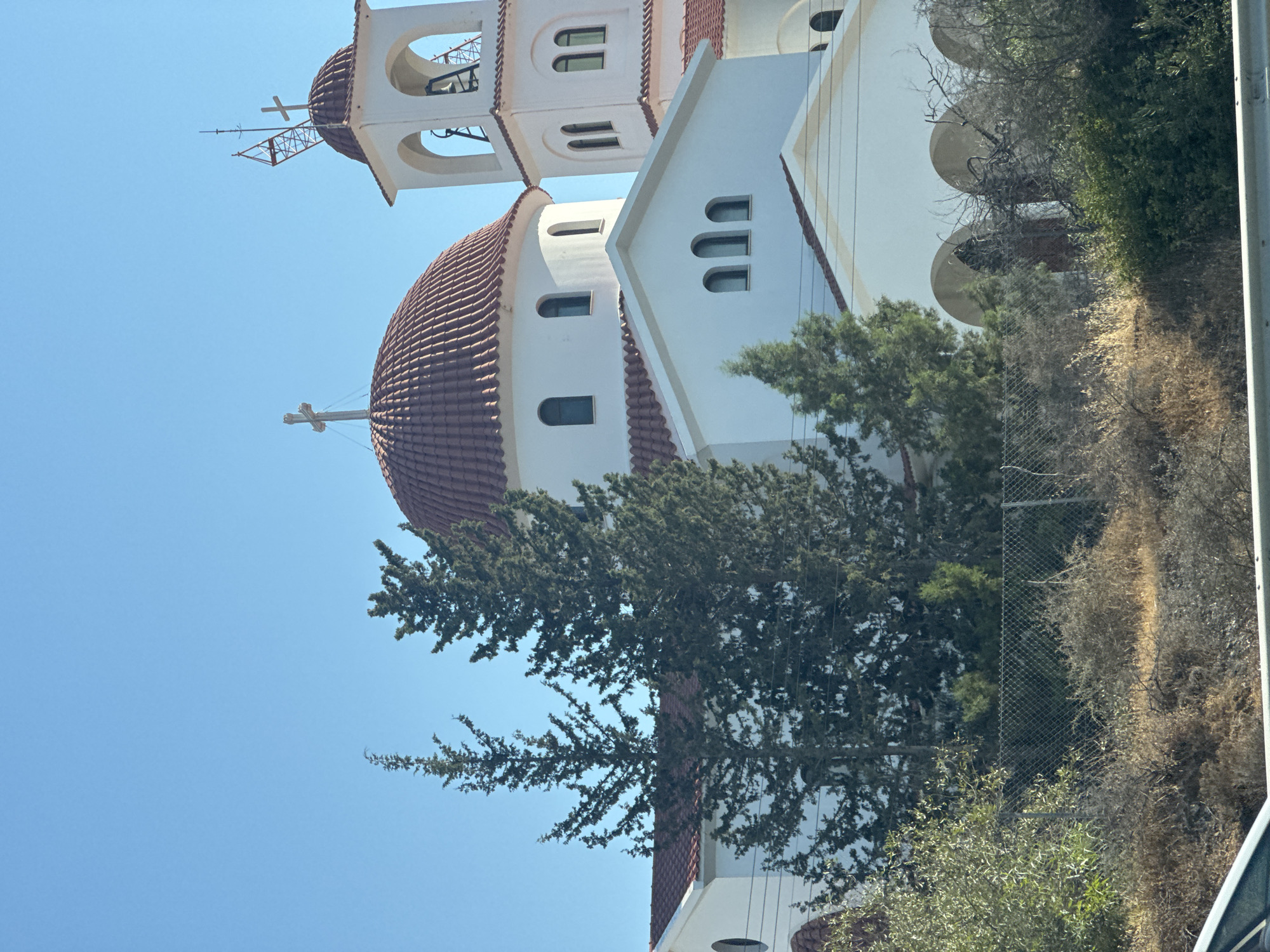 A bright, sunny day illuminates a white, domed church with a red-tiled roof and a bell tower. The church is situated in Germasogeia Municipality, Cyprus, and is surrounded by greenery and dry brush. Several tall, coniferous trees stand in front of the church, partially obscuring its facade. The sky is a clear, vibrant blue, and the overall scene is tranquil and picturesque. There are no people or animals visible in the image. The architecture of the church is traditional, with arched windows and a prominent cross atop its main dome and bell tower. The bell tower also features a construction crane-like structure.