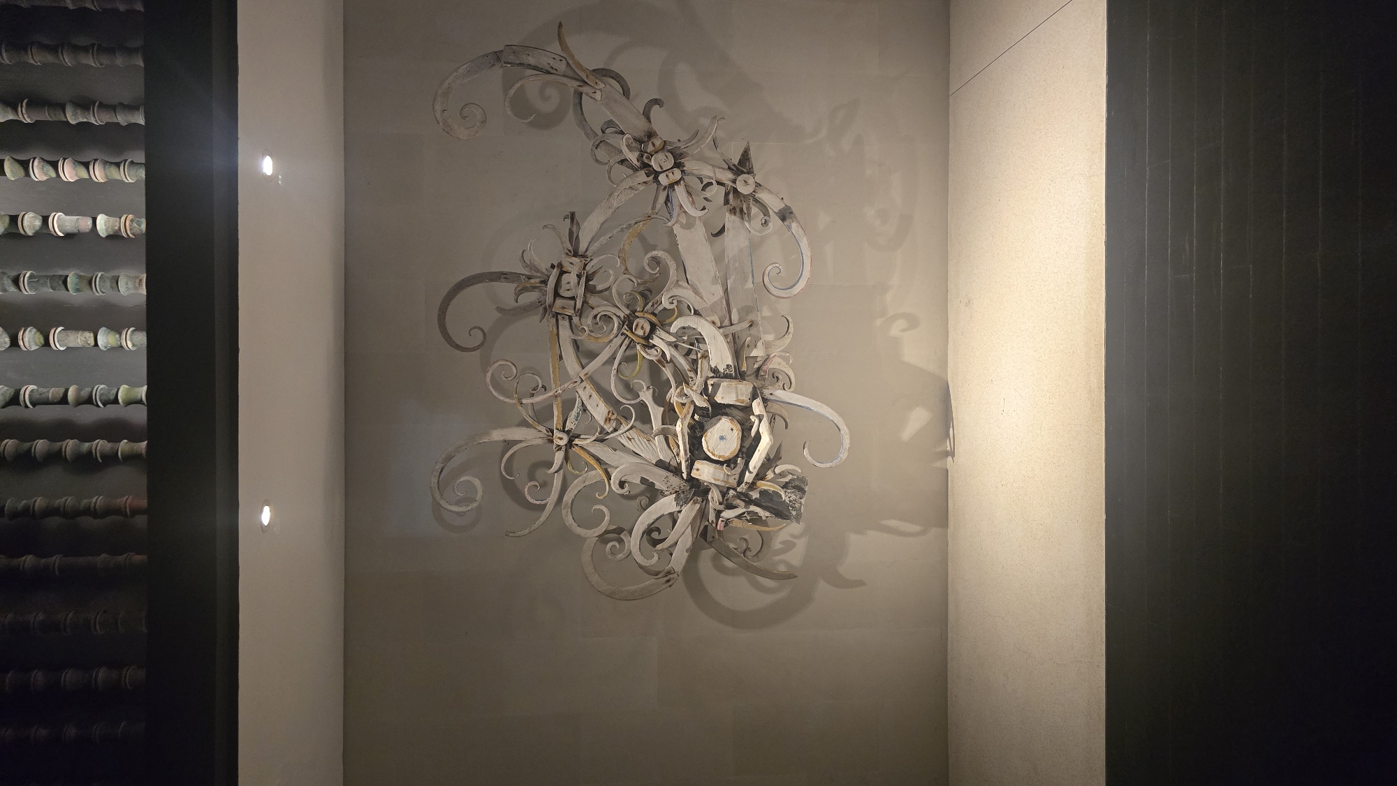 The image captures an interior space in Kuta Selatan, Indonesia. On the left, a dark structure with rows of cylindrical objects resembling bamboo or wooden poles is visible. Next to it, a narrow, light-colored wall features two small spotlights. The central focus is a decorative metal wall sculpture with floral and scrollwork designs, casting a shadow on the light-colored wall behind it. To the right, another wall, dark and textured, possibly wood or stone, extends into the frame, creating a sense of enclosure. The overall lighting is subdued, emphasizing the textures and forms within the space. No people or specific actions are depicted in the scene.