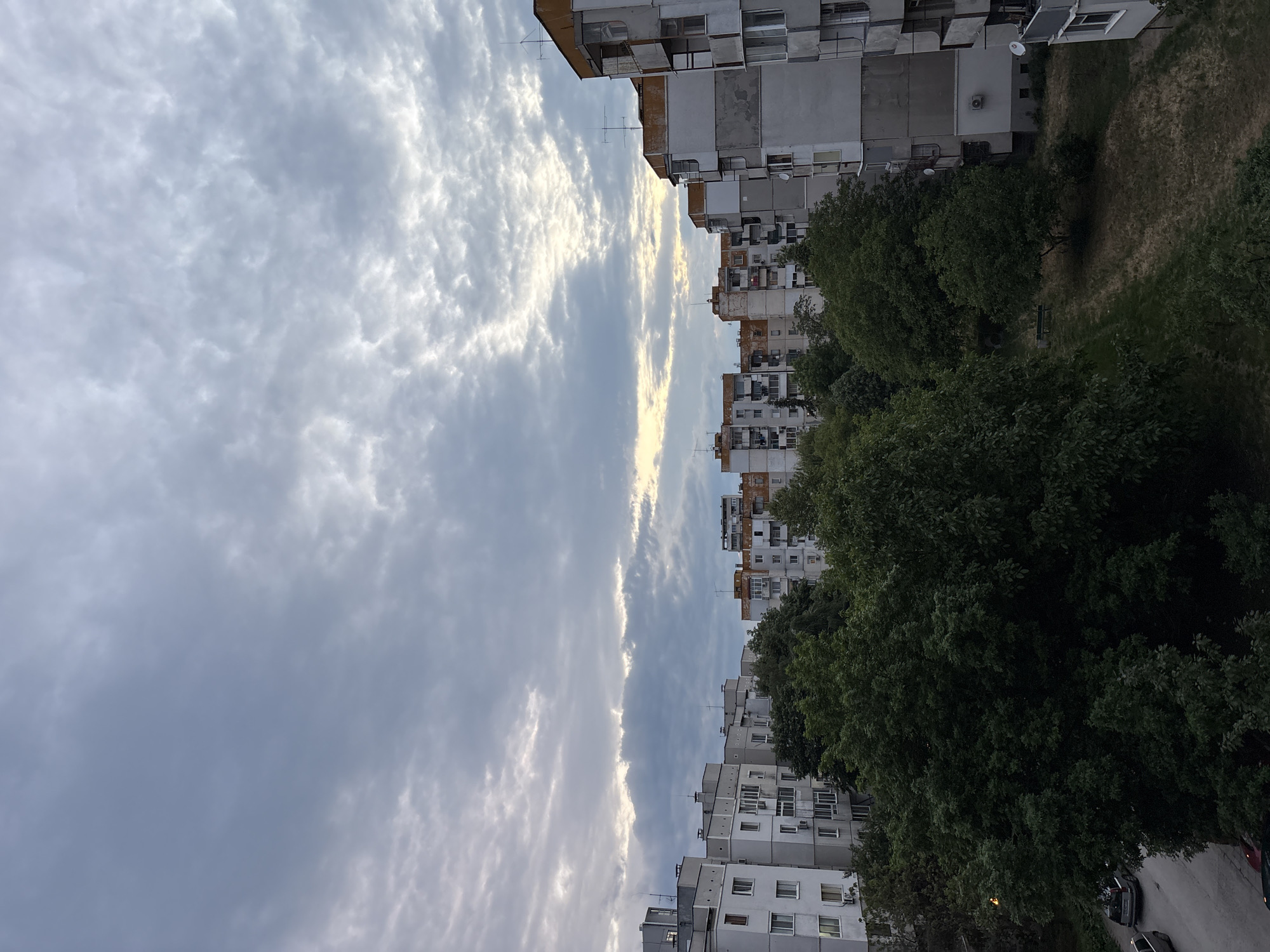 The image shows a view from an elevated position in Plovdiv, Bulgaria. The scene is divided into three main sections: the sky, buildings, and trees. The sky is overcast with clouds, featuring varying shades of gray and white. The clouds appear dense, with some lighter patches towards the horizon.
On the right side of the image are residential buildings of varying heights. They are built in a utilitarian style typical of Eastern European architecture, featuring multiple floors and balconies. The buildings show signs of aging, with visible wear and tear on their facades. There are trees growing among the buildings and along the streets below. These trees appear to be mature and lush, with full canopies that partially obscure the buildings.
The presence of the buildings and trees indicates an urban setting, and the architectural style suggests a location within the city of Plovdiv, Bulgaria.