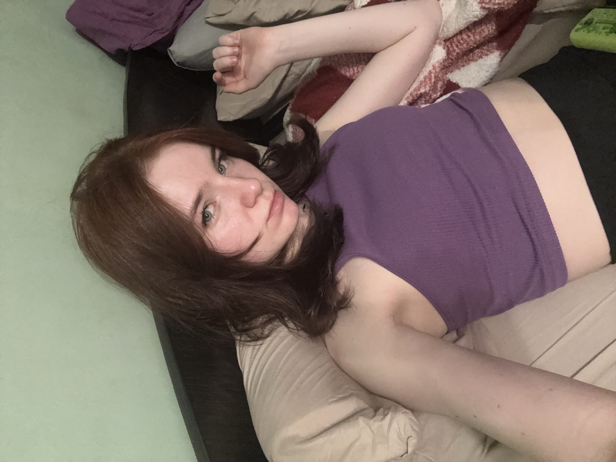 A woman is lying on a bed. She has light skin, auburn hair, and is wearing a purple top and black bottoms. She is looking directly at the camera with her head resting on a tan pillow. Behind her, there is a bed frame, a red and white patterned blanket, and a green object. The background wall is light green. The location appears to be indoors. The city and region are unknown.