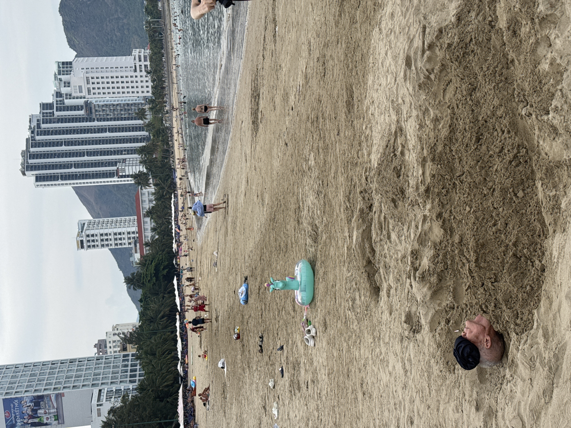 The image depicts a beach scene, likely in Bac Nha Trang Ward, Vietnam, given the distinctive high-rise buildings and mountainous background characteristic of the coastal city. The beach is crowded with people of varying ages engaged in leisure activities. Closer to the water, people are swimming and wading in the ocean. Further down the beach, some are walking, relaxing under umbrellas, or sunbathing. There's a noticeable amount of scattered personal belongings, including shoes, bags, and inflatable toys, strewn across the sand. The foreground shows a man partially buried in the sand with only his head visible, and he is wearing a black cap. The overall environment indicates a popular tourist destination.