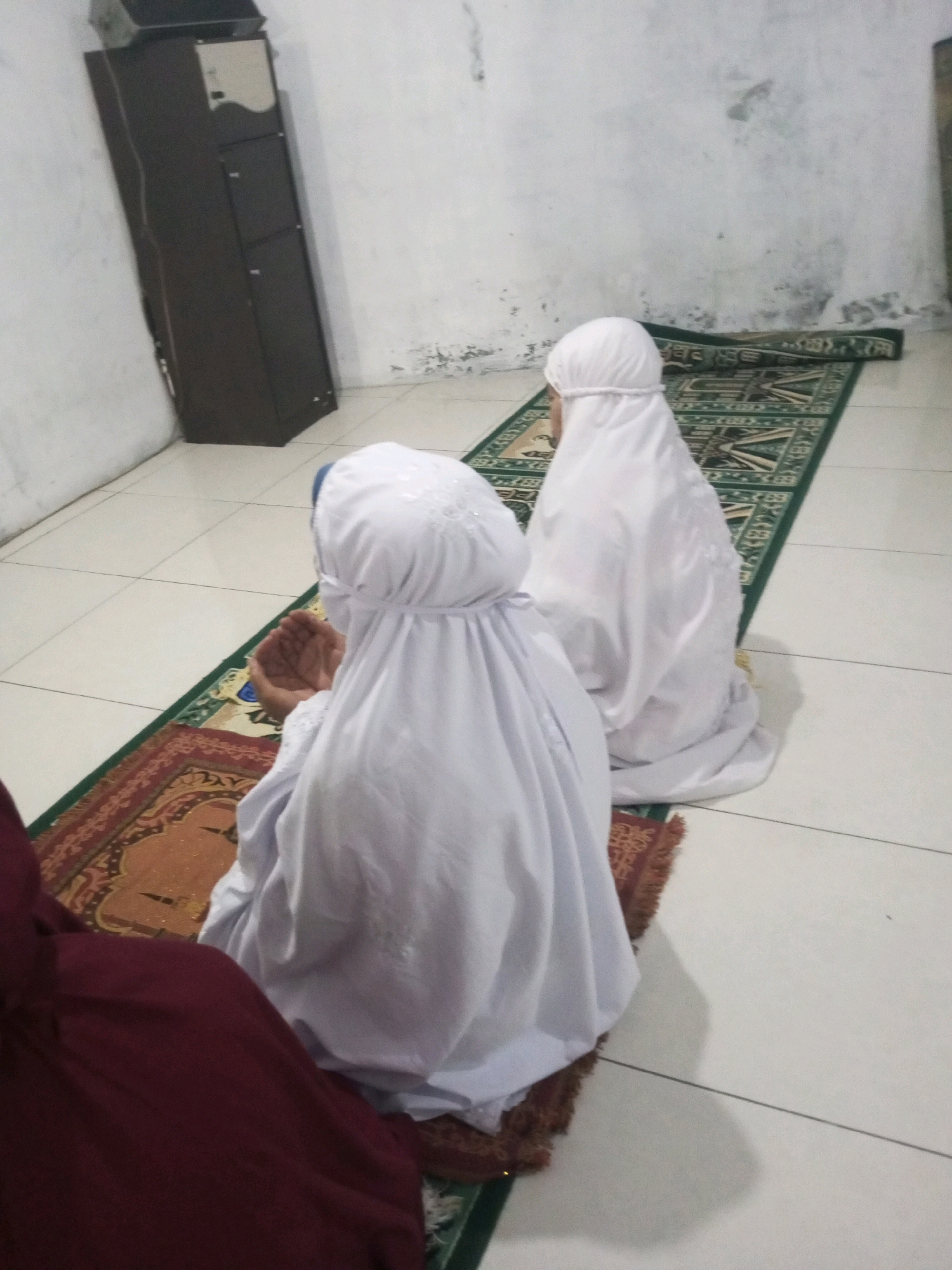 In a room with tiled flooring and textured walls, two individuals are kneeling on prayer mats. The individual in the foreground, facing away from the camera, is wearing a white headscarf and covering. Their hands are held together in prayer. To the right and slightly behind, a second individual, also in white attire, is also in a kneeling prayer posture. A tall, dark wooden cabinet stands against the left wall, with a lighter colored section at the top. The prayer mats have intricate patterns. This scene depicts an act of worship within a domestic or communal space.