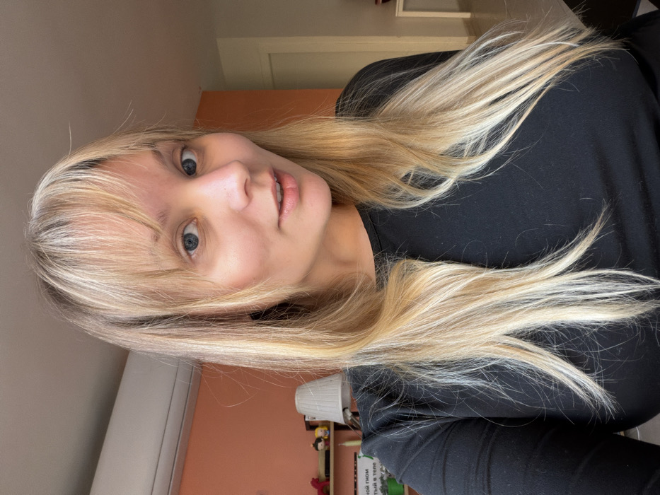 The image displays a head and upper torso portrait of a fair-skinned female subject, approximately in her late 20s to early 30s, situated indoors within what appears to be a residential setting in Tel-Aviv, Israel.
The subject has long, straight to slightly wavy blonde hair with lighter highlights, parted centrally with a full fringe covering her forehead. Her eyes are blue and directed toward the camera. She exhibits a subtle, open-mouthed smile. She is wearing a plain, long-sleeved black top. No visible jewelry or tattoos are present.
The background reveals a room with a peach-orange painted wall on the left side, transitioning to a lighter, possibly off-white, color on the right. A white split-unit air conditioning system is mounted high on the peach-orange wall in the upper left quadrant. Below the air conditioner, a light-colored shelf or ledge is visible, hosting several small items including what appears to be a red figurine, two small collectible figures, and a white table lamp with a pleated shade. Below the lamp, a white rectangular object displays Russian text, partially visible, which reads "НОЙ ГНОМ" on the top line and "ТЫЙ В ТЕЛЕ" on the second line. Further down, green potted plants are partially visible. To the right, a doorway with a light-colored frame leads to an adjacent room, where a white table is partially visible on the far right edge of the frame. Natural light appears to illuminate the scene, primarily from the right side.