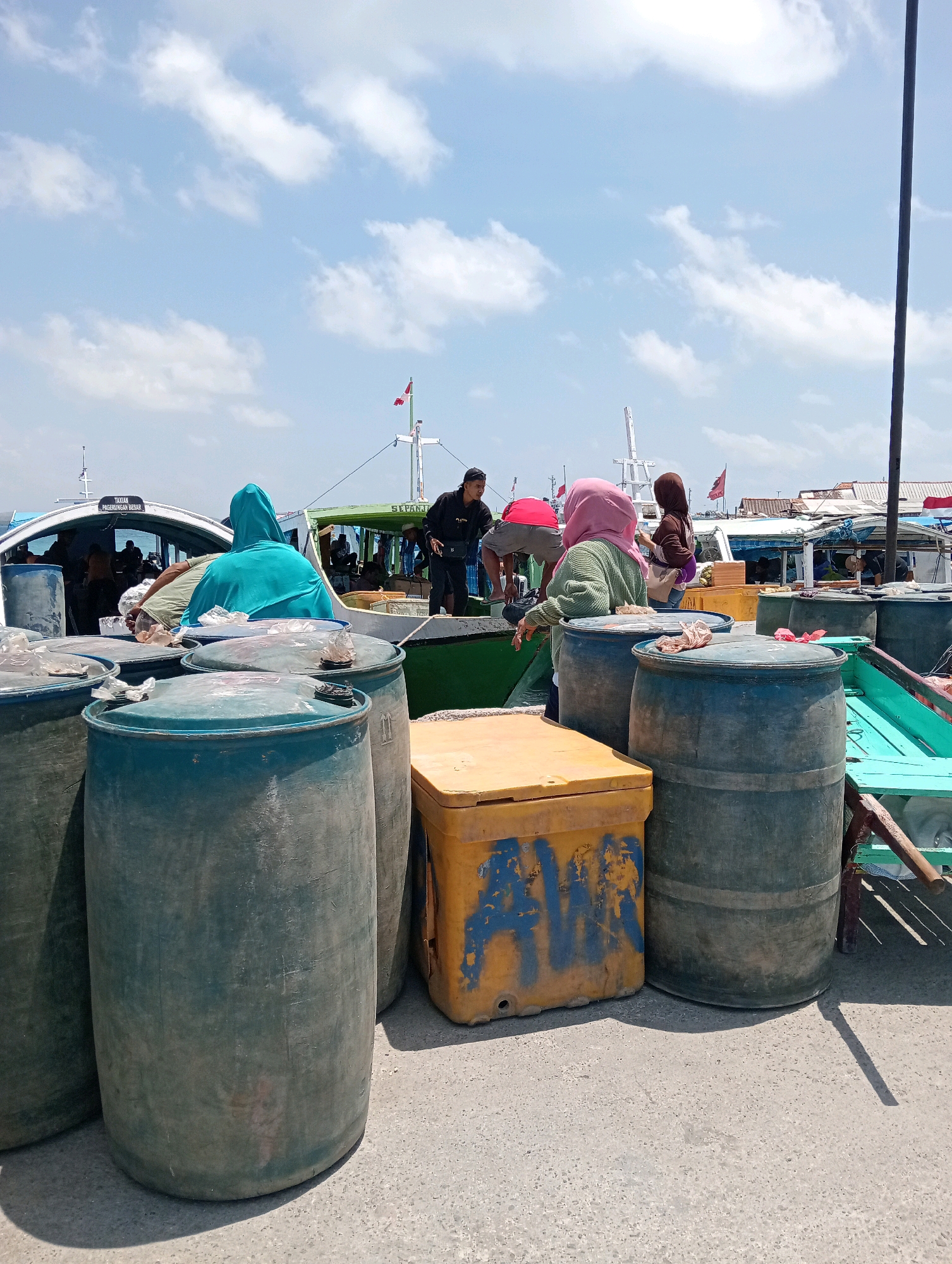 The image depicts a scene at the port of Sapeken, Indonesia, under a bright, partially cloudy sky. In the foreground, several large, dark green plastic barrels are prominent, some with lids partially covered by cloth. To the right of the barrels, a yellow cooler with blue graffiti is visible. Behind these objects, a green boat with people on board is docked. Several individuals are seen interacting on and around the boat. Some are wearing headscarves. The background reveals more boats and structures typical of a harbor environment. The ground is paved with asphalt or concrete. The overall impression is one of a busy working port.
