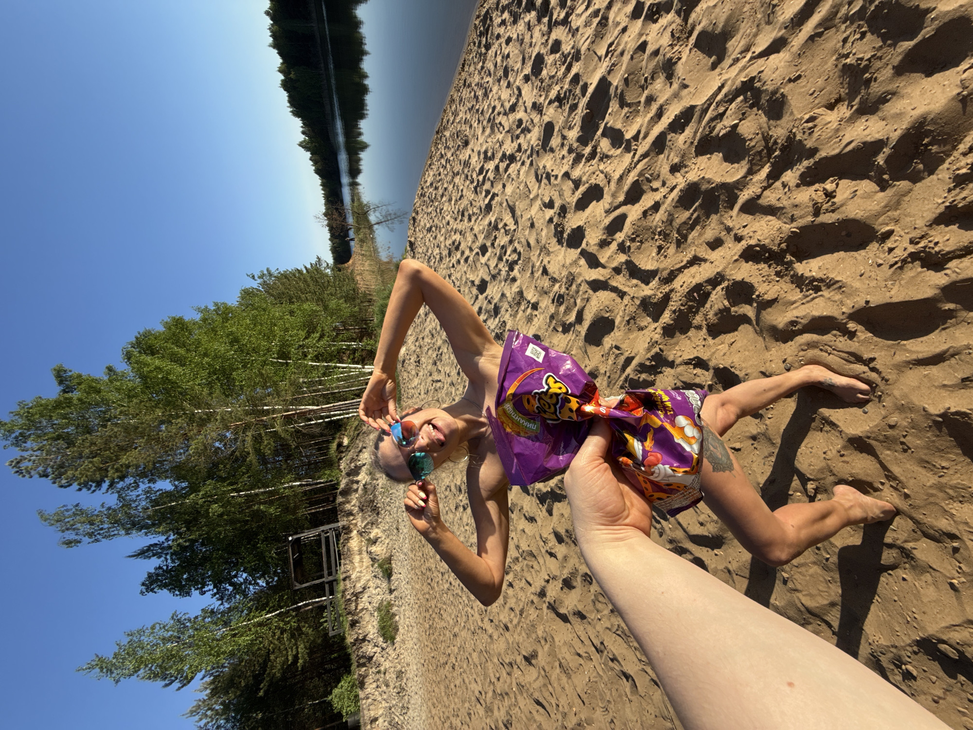 A person is lying on a sandy beach next to a body of water. The person is wearing sunglasses and holding them, and they are holding a purple bag of potato chips over their body. A person's arm reaches into the frame, holding the potato chips. The location is likely a beach near Novyi, Russia. The sky is blue and sunny. Trees are visible to the left.