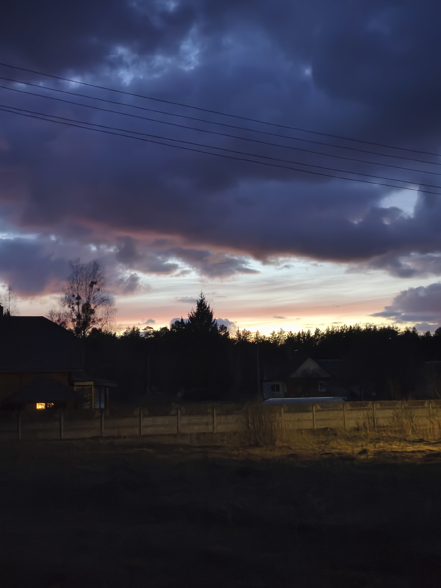 The image depicts a rural scene in Парэчча, Belarus at dusk. In the foreground, a dark field is visible. Behind it stands a weathered, concrete fence illuminated by a faint, diffuse light source, potentially a street lamp. Behind the fence are houses and a dense line of trees silhouetted against the colorful sunset sky. The sky is a mix of dark, stormy clouds in shades of purple and blue, with a band of orange and pink sunlight peeking through near the horizon. Power lines stretch across the upper part of the frame. A single illuminated window is visible in one of the houses. A bare tree with prominent mistletoe clumps stands near the left side of the image.