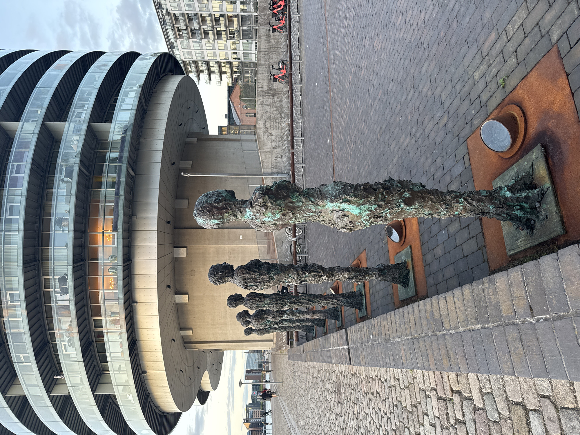 This is a medium-shot, eye-level, outdoor photograph taken at dusk in Copenhagen, Denmark. In the foreground, a line of seven bronze statues of human figures stands on a paved walkway. The statues are textured and appear weathered, with a greenish patina. They are positioned on square, rust-colored bases. The walkway itself is paved with dark gray bricks, with a cobblestone path to the left. In the background, a large, modern, multi-story building with curved balconies and glass windows dominates the scene. To the right of this building, a lighter-colored apartment building with multiple balconies is visible. A row of parked bicycles and what appear to be electric scooters are also present in the middle ground. The sky is overcast with a soft, diffused light. The overall atmosphere is calm and serene, with the statues creating a sense of human presence against the backdrop of urban architecture.