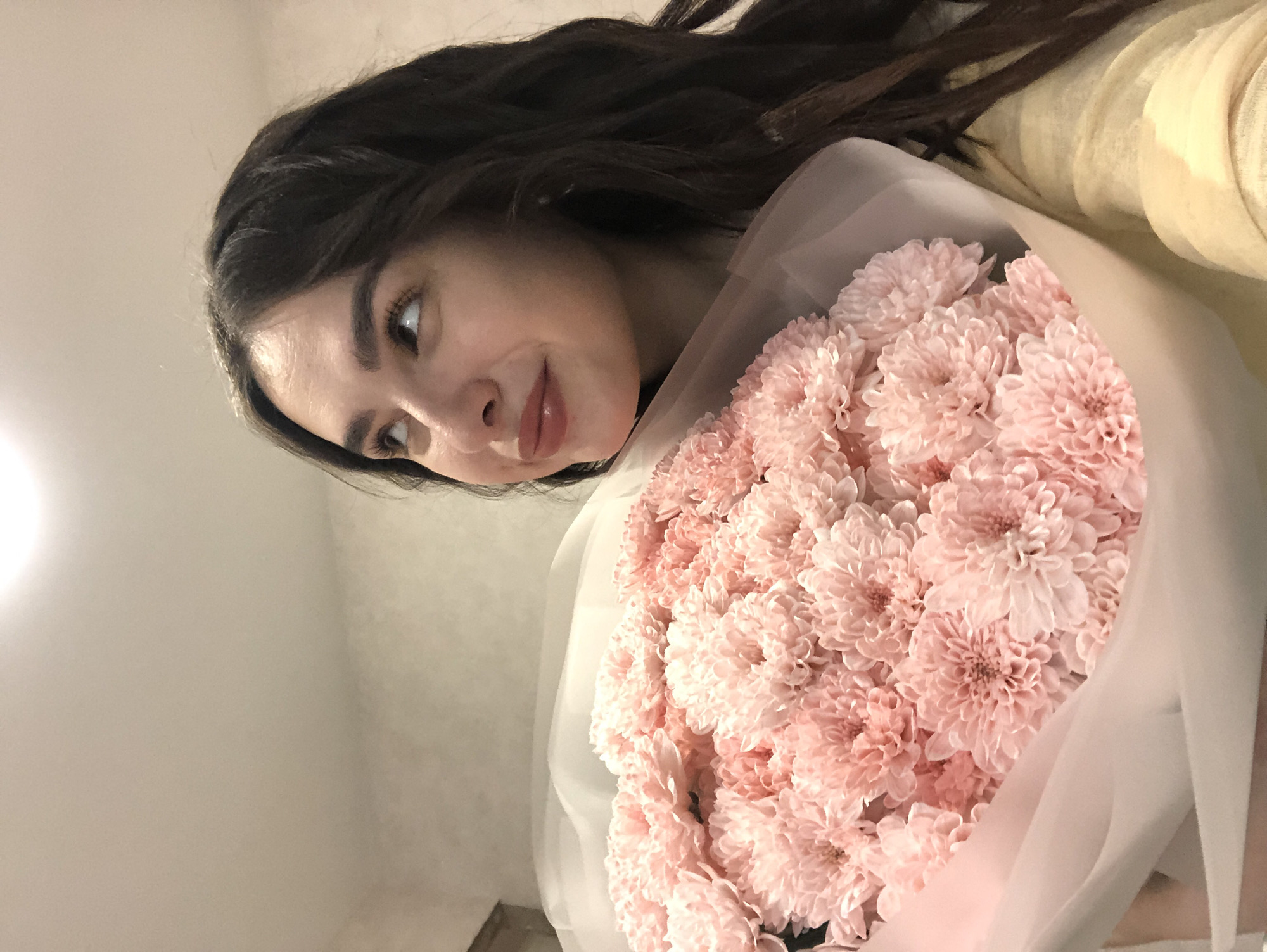 A young woman with dark, wavy hair is holding a large bouquet of light pink chrysanthemums. She is looking upwards and to her right with a gentle smile, her lips slightly parted. Her skin is fair, and she has dark eyebrows and eyelashes. She is wearing a light yellow top. The bouquet is wrapped in sheer white and pink paper. The background is a plain, lightly textured wall with a bright light source visible at the top. The scene is indoors. There is no information in the image to indicate the location in Chaikovsky, Russia.