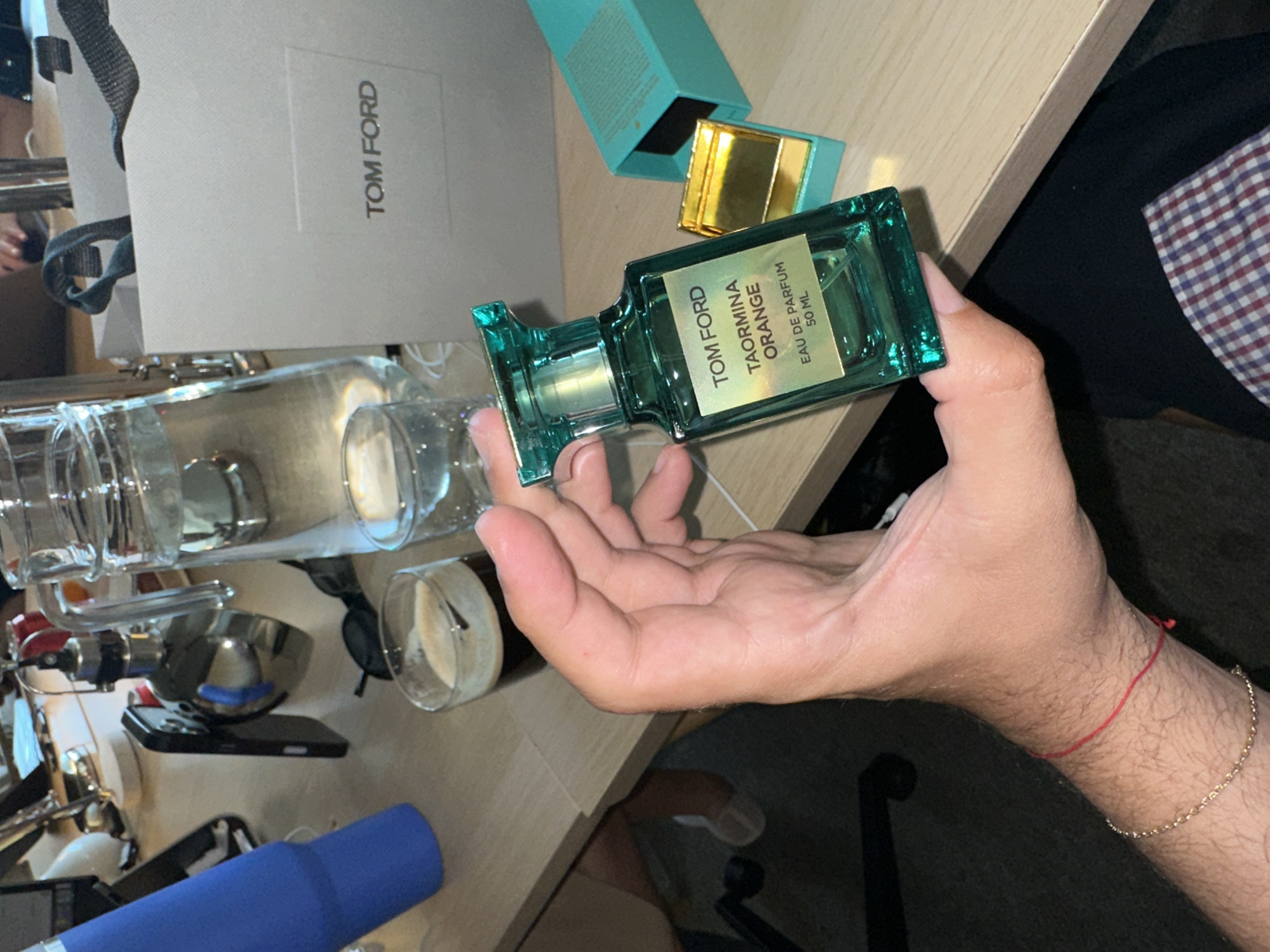 A hand is holding a Tom Ford Taormina Orange perfume bottle. Nearby are the box for the perfume, a Tom Ford shopping bag, glasses, and personal items on a desk. The location is Unknown, Unknown.