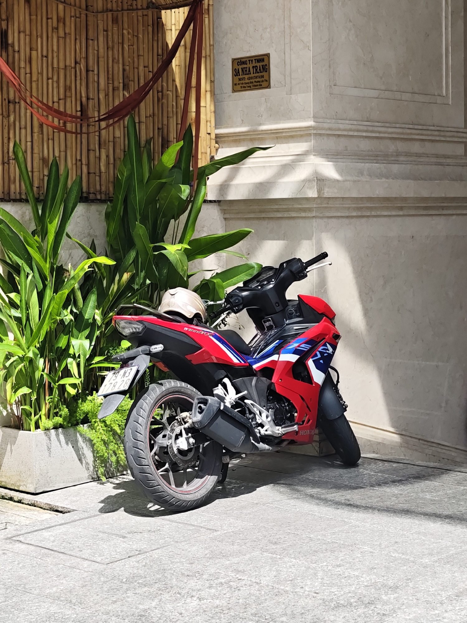 In Nha Trang Ward, Vietnam, a red and blue motorcycle is parked on a paved sidewalk next to a planter filled with lush green foliage. A light-colored helmet rests on the rear seat of the motorcycle. Behind the motorcycle and planter, a wall with a textured finish is visible, adorned with a sign that reads "CÔNG TY TNHHH 3A NHA TRANG" and includes other Vietnamese text. A bamboo screen with red ribbons hangs on the left side of the scene. The ground is paved with square tiles, and shadows from the motorcycle and plants are cast on the pavement.
