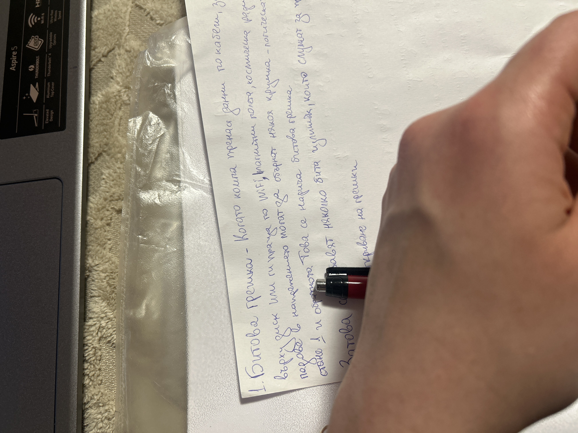 A hand is holding a mechanical pencil and writing on a sheet of white paper. The paper has text in Bulgarian, beginning with "1. Битова грешка". The text describes bit errors and their causes. To the left of the paper is a gray laptop with the text "Aspire 5" visible. The laptop features various logos indicating Wi-Fi 6, Thunderbolt 4, and SSD capabilities. The laptop rests on a beige surface that appears to be a towel or similar textile. The scene does not provide enough information to determine its location in Burgas, Bulgaria.