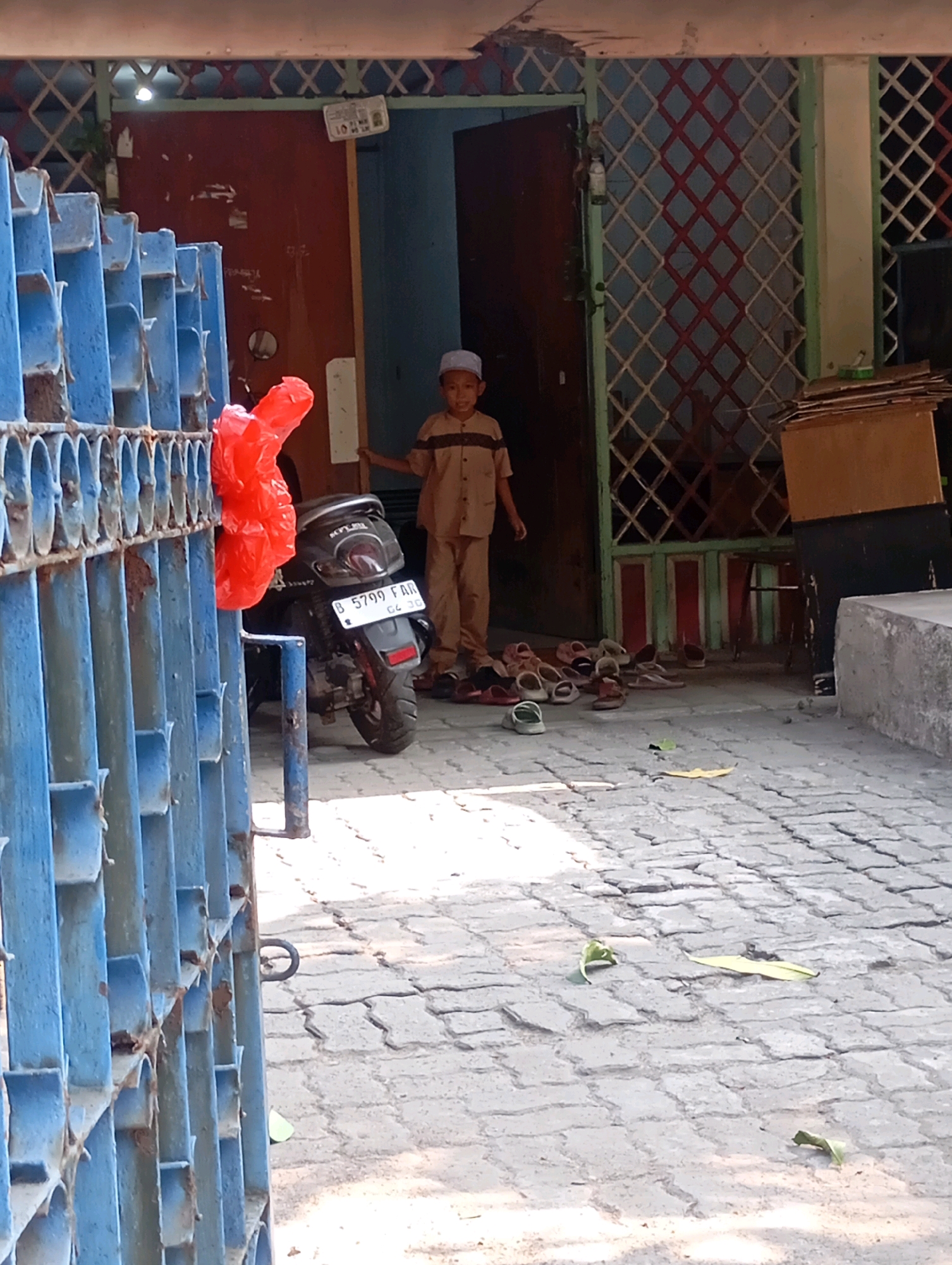 The image depicts an outdoor scene in front of a building, likely a mosque or a community center, in Babelan, Indonesia. A young boy wearing a light brown collared shirt and matching pants, along with a white skullcap, stands near the entrance, holding a wooden stick with a sign attached to it. In the foreground, a blue metal gate with vertical bars is visible. Behind the gate, a motorcycle with a red plastic bag attached to its handlebars is parked. The ground is paved with interlocking concrete blocks, and several pairs of sandals are scattered near the building's entrance. The building's facade features a red door and a window with a diamond-patterned grille. Sunlight casts shadows on the ground, indicating it is daytime. The overall impression is a snapshot of daily life in a neighborhood setting.