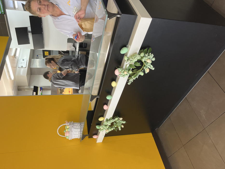 A black counter with a glass top is decorated with a white shelf. There are green and pink Easter eggs and green plant wreaths on the shelf. The counter is in a fast food restaurant, and two people are working behind it, while a third person stands beside the counter, holding a brown paper bag in their hand. The restaurant is located in Hadjidimitrovo, Bulgaria. The walls are yellow. A white wicker basket with pink and green Easter eggs inside rests on top of the counter.