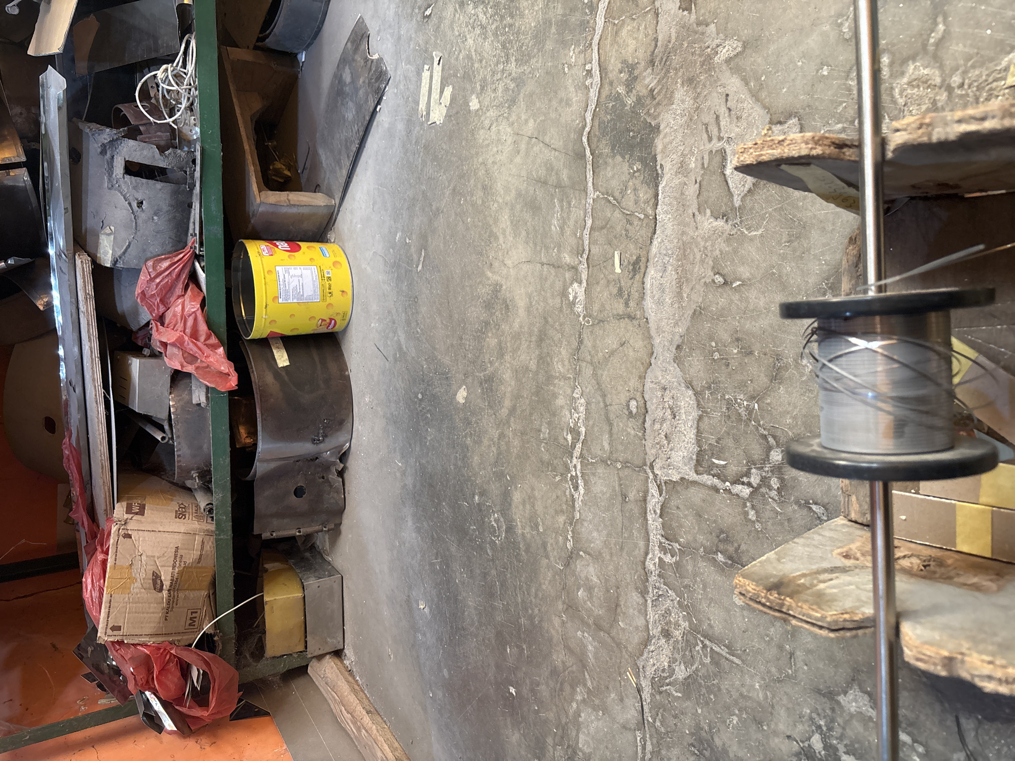The image depicts a cluttered workshop or storage area, likely in Telagamurni, Indonesia. In the foreground, a spool of grey wire is mounted on a wooden apparatus. Behind this, shelves are stacked with various items, including red plastic bags, cardboard boxes, metal parts, and a yellow cylindrical container. The floor is concrete, showing signs of wear and tear with cracks and debris. The overall impression is one of an organized but well-used space, possibly for mechanical work or storage of industrial materials.