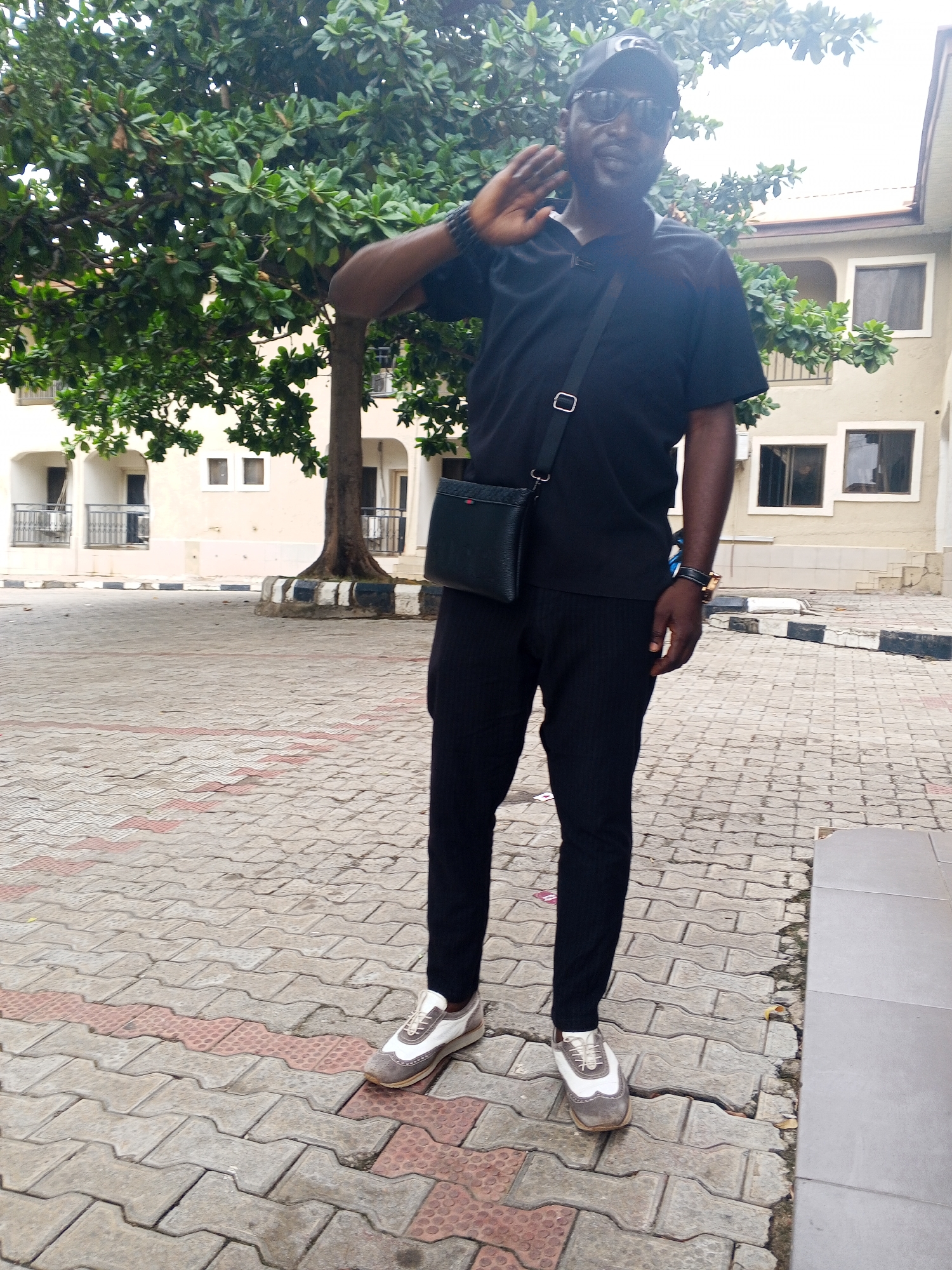 A man stands outdoors on a paved surface. He is wearing a black t-shirt, black pants, a black cap, and sunglasses. A black bag hangs across his shoulder. His left hand is raised, as if waving or saluting. Behind him is a building with multiple windows and balconies and a leafy tree. The location is Lokoja, Nigeria.