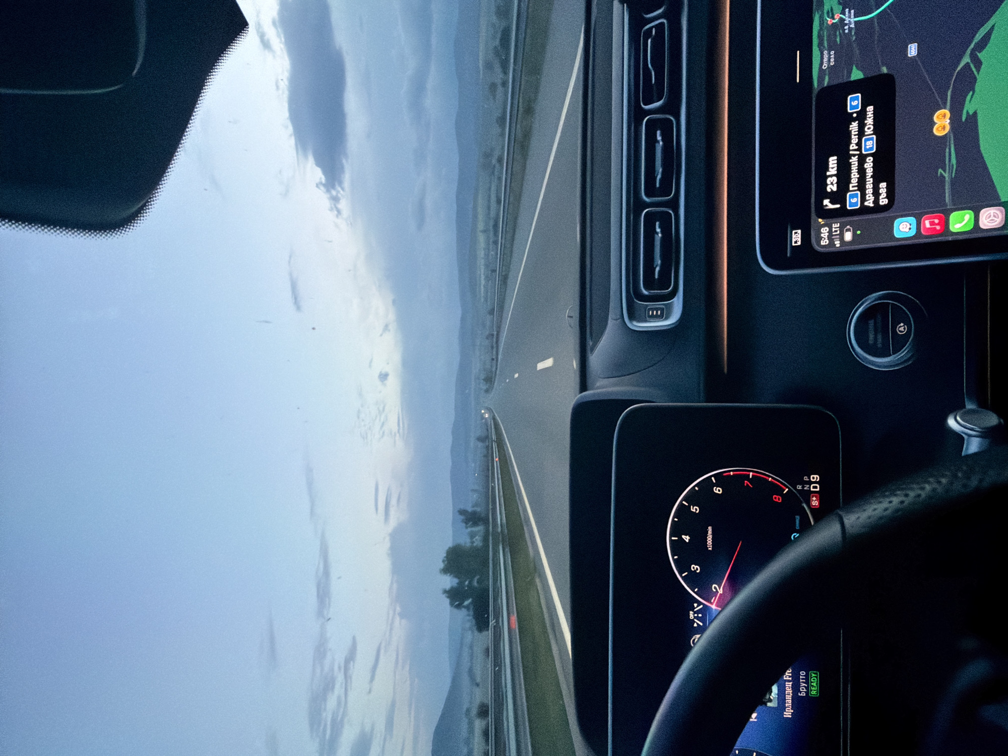 The image shows the interior of a car, likely a Mercedes-Benz, driving on a highway. The dashboard is visible, with the instrument panel showing the tachometer and a media display. A smartphone mounted on the dashboard displays navigation information indicating that the car is approximately 23 km away from Pernik, Bulgaria, specifically the Draghichevo area along the "South Arc". The road ahead appears to be a multi-lane highway with a median. The sky is overcast. The steering wheel and portions of the car's interior, including air vents, are also visible. The display says the car is playing an Irish music called Брутто ("Brutto").