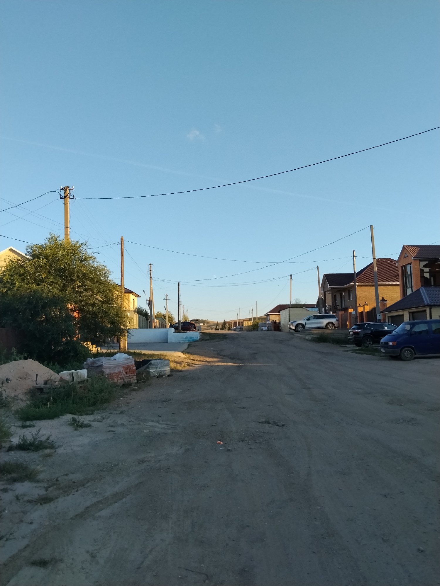 The image depicts a dusty, unpaved road in a residential area, likely a suburban or rural part of Kokshetau, Kazakhstan. The scene is characterized by several houses, a large green tree on the left, and utility poles with overhead wires. Construction materials, including piles of sand and bricks, are visible on the left side of the road, suggesting ongoing development or renovation. Several vehicles, including a blue van, a black SUV, and a white SUV, are parked along the right side of the road. The sky is clear and blue with a faint cloud streak. The overall impression is one of a developing community under a bright sky.