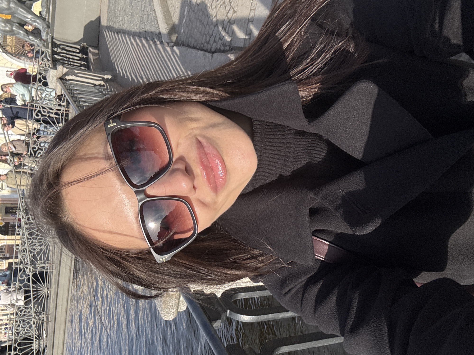 A woman is taking a selfie outdoors, likely in Saint Petersburg, Russia. She is wearing sunglasses and a black coat. In the background, there is a body of water and an ornate metal fence behind which many people are visible, suggesting a popular tourist spot. The image is slightly tilted.