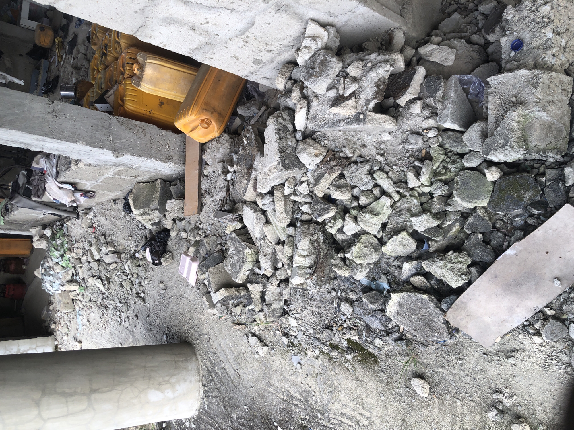 This image depicts a scene of debris and construction materials in Ado, Nigeria. A large, grey concrete pillar dominates the left side of the frame. The ground is covered with rubble, including broken concrete pieces, dirt, and scattered debris. In the background, several yellow plastic jerrycans are stacked, suggesting they may contain liquids. Some pieces of fabric and other miscellaneous items are also visible amidst the rubble. The environment appears to be an outdoor construction or demolition site, characterized by a cluttered and unkempt appearance. There are no people visible in the image.