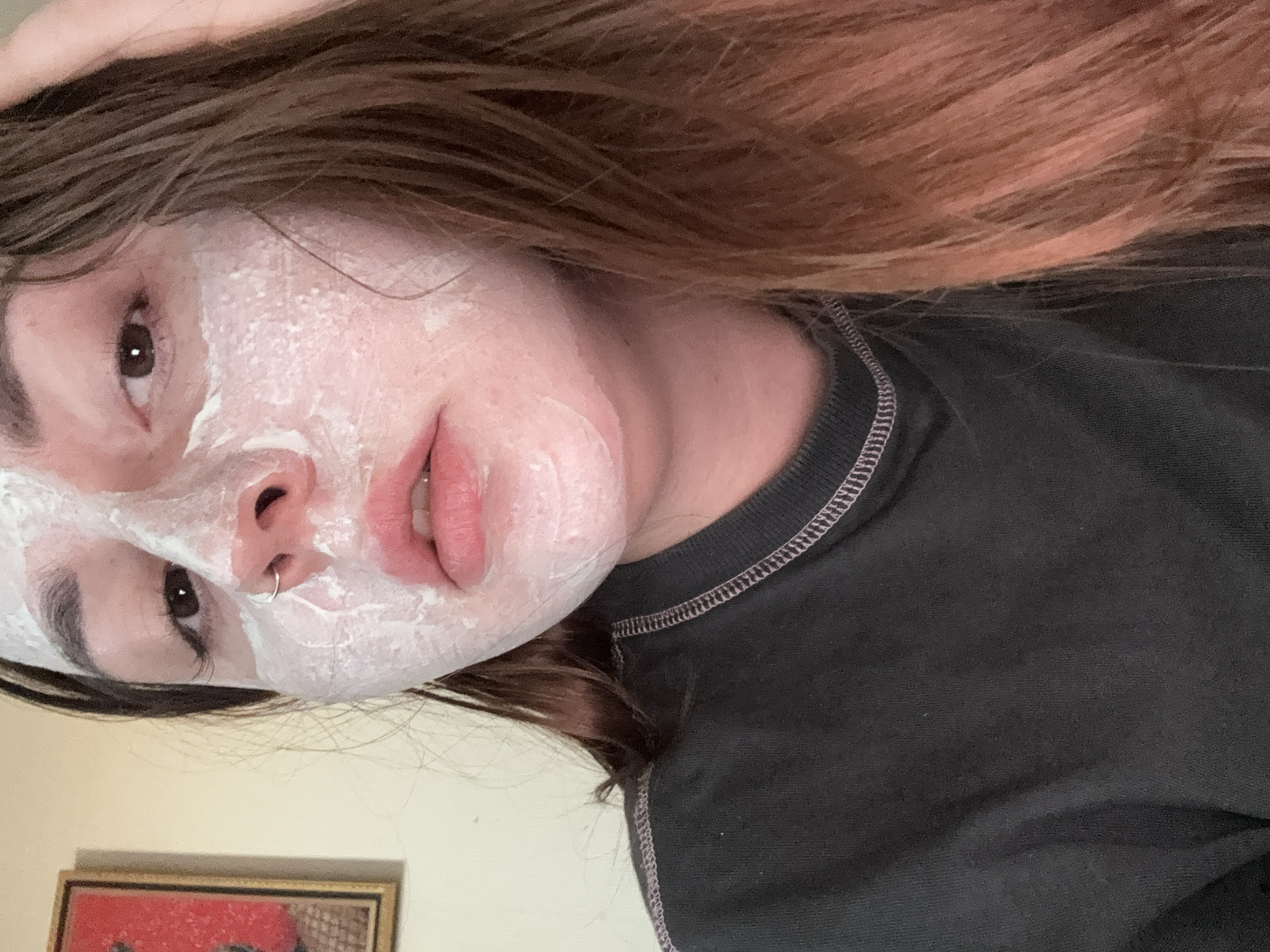A close-up selfie depicts a young woman with a white facial mask. Her hair is brown with reddish highlights. She is wearing a dark grey top with a stitched neckline. She has a nose ring in her left nostril. The background is out of focus, but includes a framed picture with red visible in the corner. There is no indication that the location is Izhevsk, Russia.