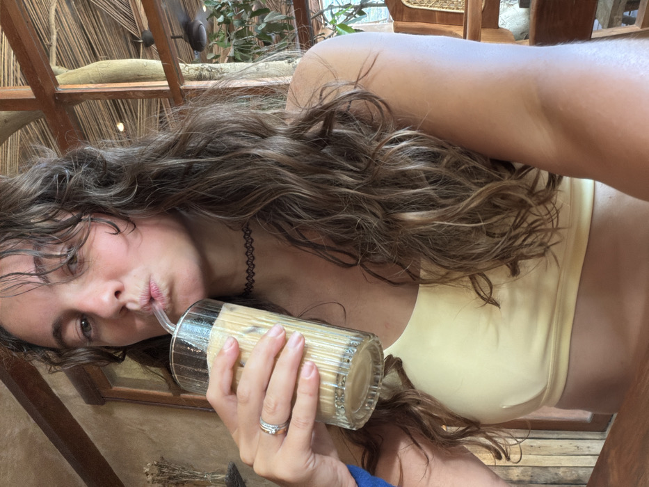 A woman reclines in a yellow top, sipping from a glass of iced coffee with a straw. She wears a delicate choker and a ring on her finger. Her long, wavy hair cascades around her face and shoulders. The background features a textured, possibly thatched, structure. A glimpse of greenery is visible in the top center, possibly indicating an outdoor or semi-outdoor setting.