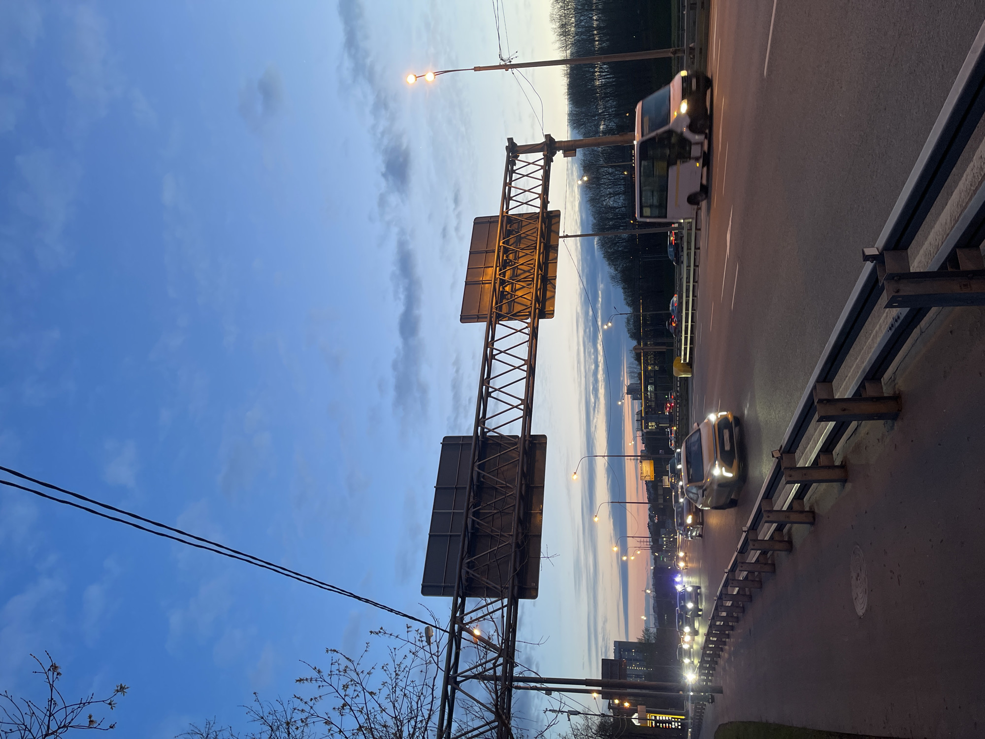 This landscape photograph captures an urban highway scene at dusk, likely in Moscow, Russia. The sky dominates the upper portion of the frame, displaying a gradient from deep blue at the top to lighter blues and soft orange/pink hues near the horizon, characteristic of twilight. Scattered clouds are visible throughout the sky.
The main subject is a multi-lane road or overpass, with several vehicles in motion. Car headlights are illuminated, casting light on the asphalt. A white minibus is prominent, along with a light-colored sedan (possibly gold or yellow). To the right, a metal guardrail runs parallel to the road.
A large, metallic overhead road sign structure, seen from its rear side with a visible latticework support, spans across the highway. Numerous streetlights, some brightly lit, line the road, contributing to the evening illumination. Power lines traverse the upper sky. In the lower left, bare tree branches with visible budding leaves indicate early spring. The distant background features a dark silhouette of trees and faint outlines of urban buildings, confirming the city setting. There is no legible text visible on the signs. The overall scene depicts typical evening traffic on an urban arterial road.