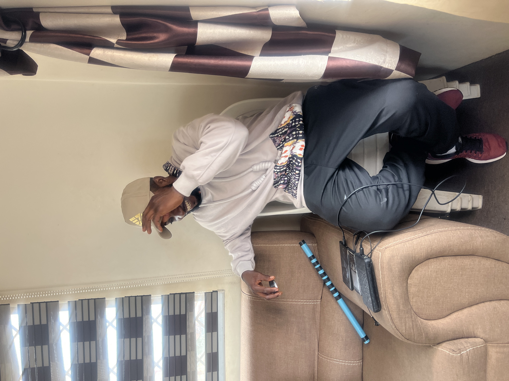 A man wearing a beige Adidas baseball cap and a light-colored hoodie is sitting on a chair next to a brown couch. He is laughing and covering his face with his hand. On the couch, there are two phones and a power bank plugged in, along with a blue and black striped cylindrical object. To the right of the man is a window with blinds and a patterned curtain hangs on the wall. The overall scene suggests a casual indoor setting, possibly a living room, in Jos, Nigeria.